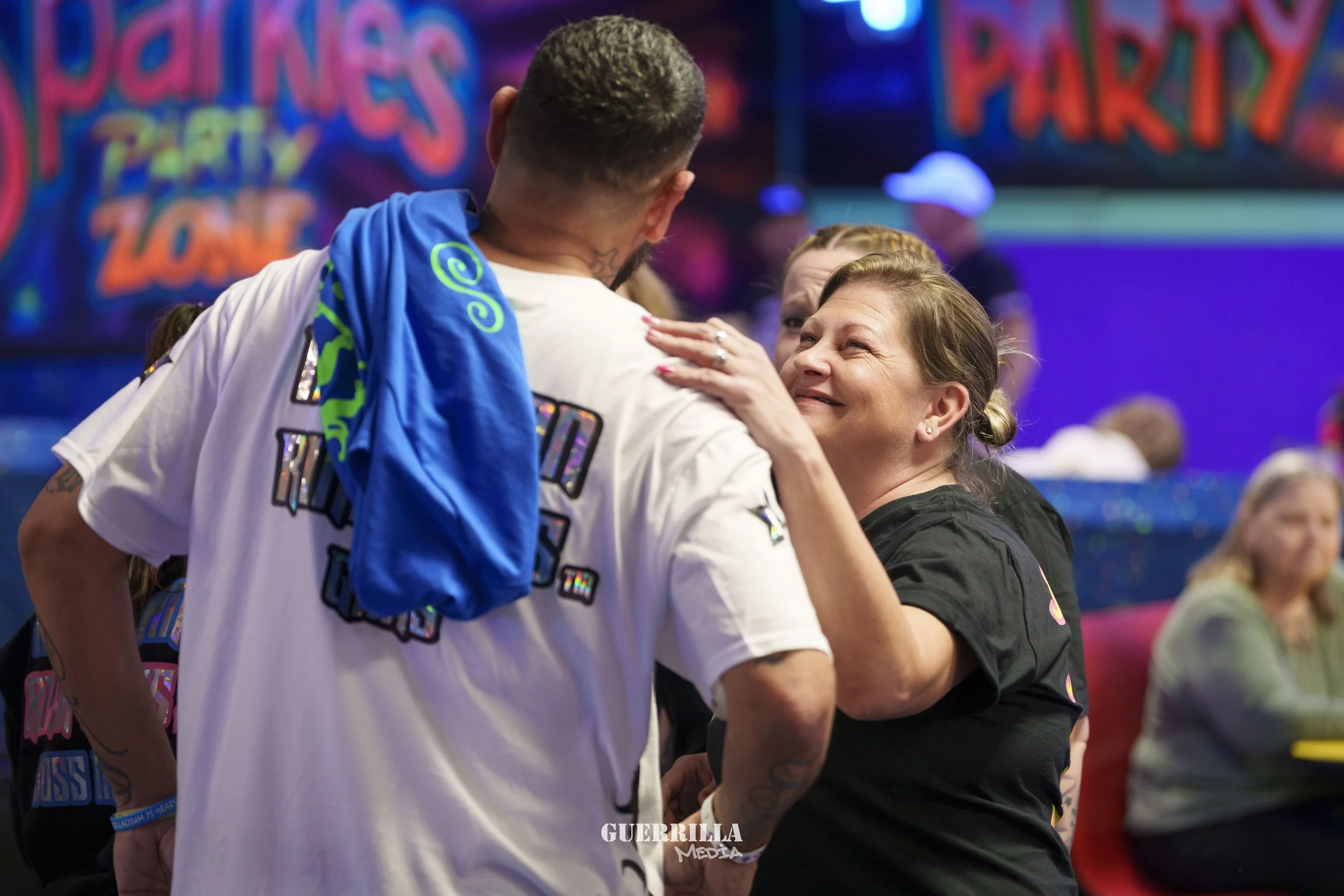 A woman embraces a man with a blue towel draped over his shoulder at a party zone. The background has colorful party signs and decorations.