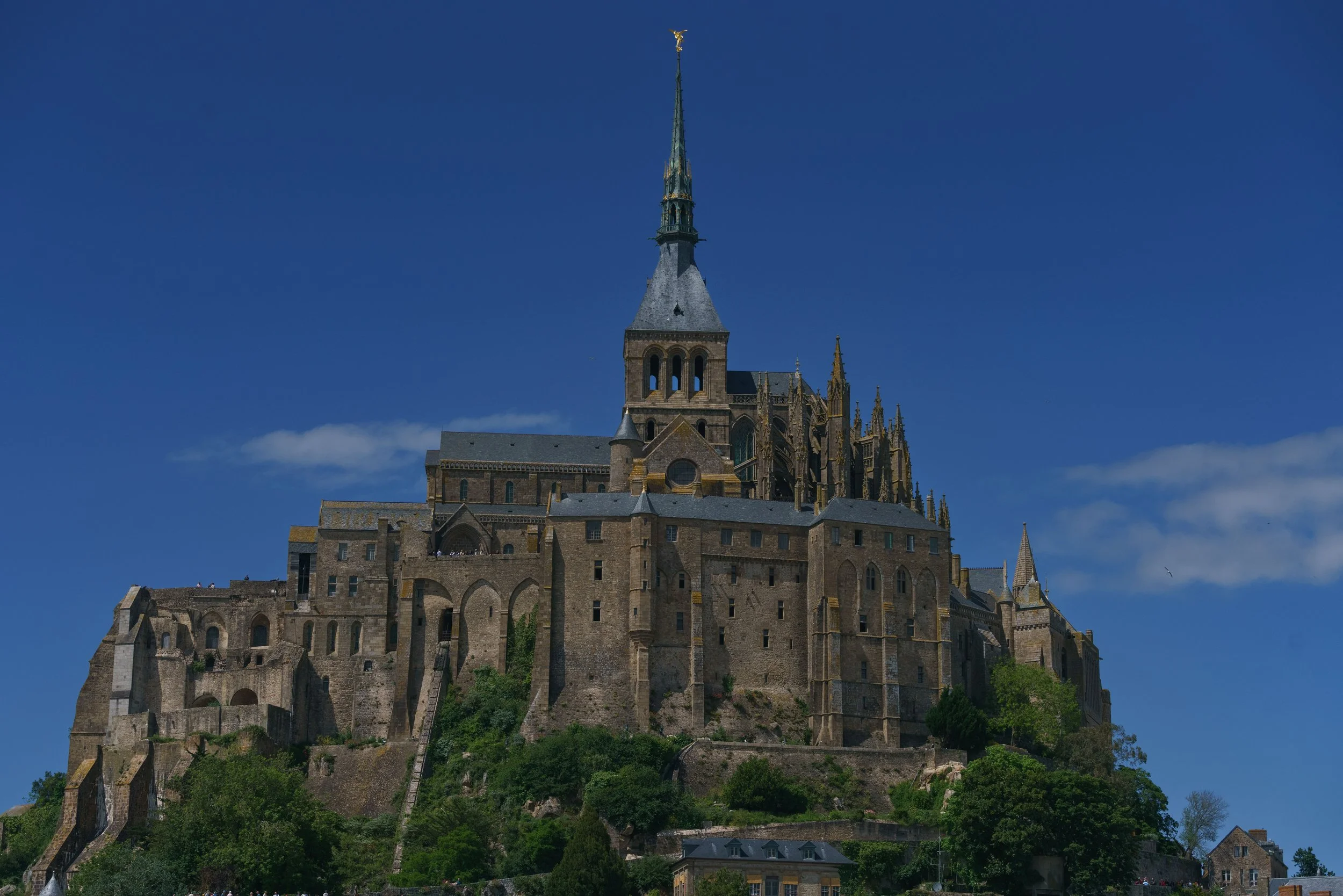 A large historic castle on a hilltop with a tall spire and Gothic architecture, surrounded by greenery and under a clear blue sky.