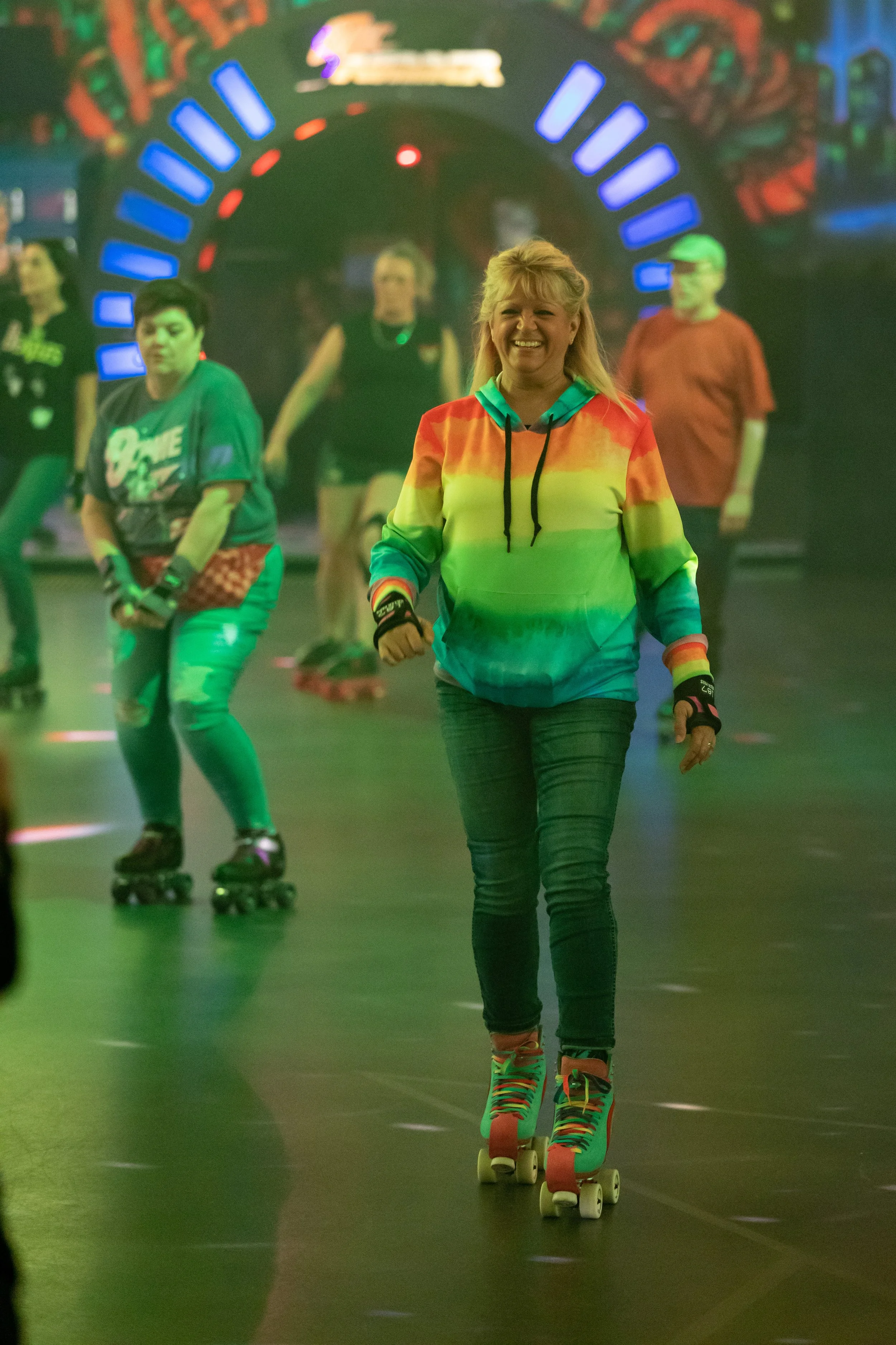 A woman roller skating indoors, smiling, wearing a rainbow-colored hoodie, black jeans, and colorful roller skates, with a group of people in the background also skating.