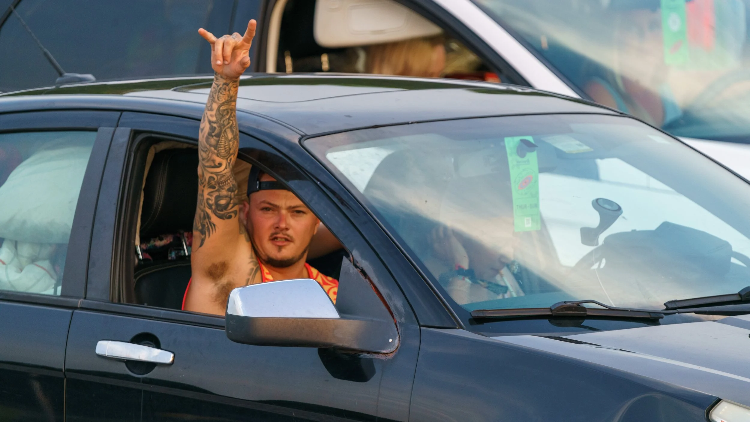 A man with tattoos on his arm is sitting in a car, raising his right arm with a gesture, looking out the window with a serious expression, wearing a backwards cap and a red shirt.