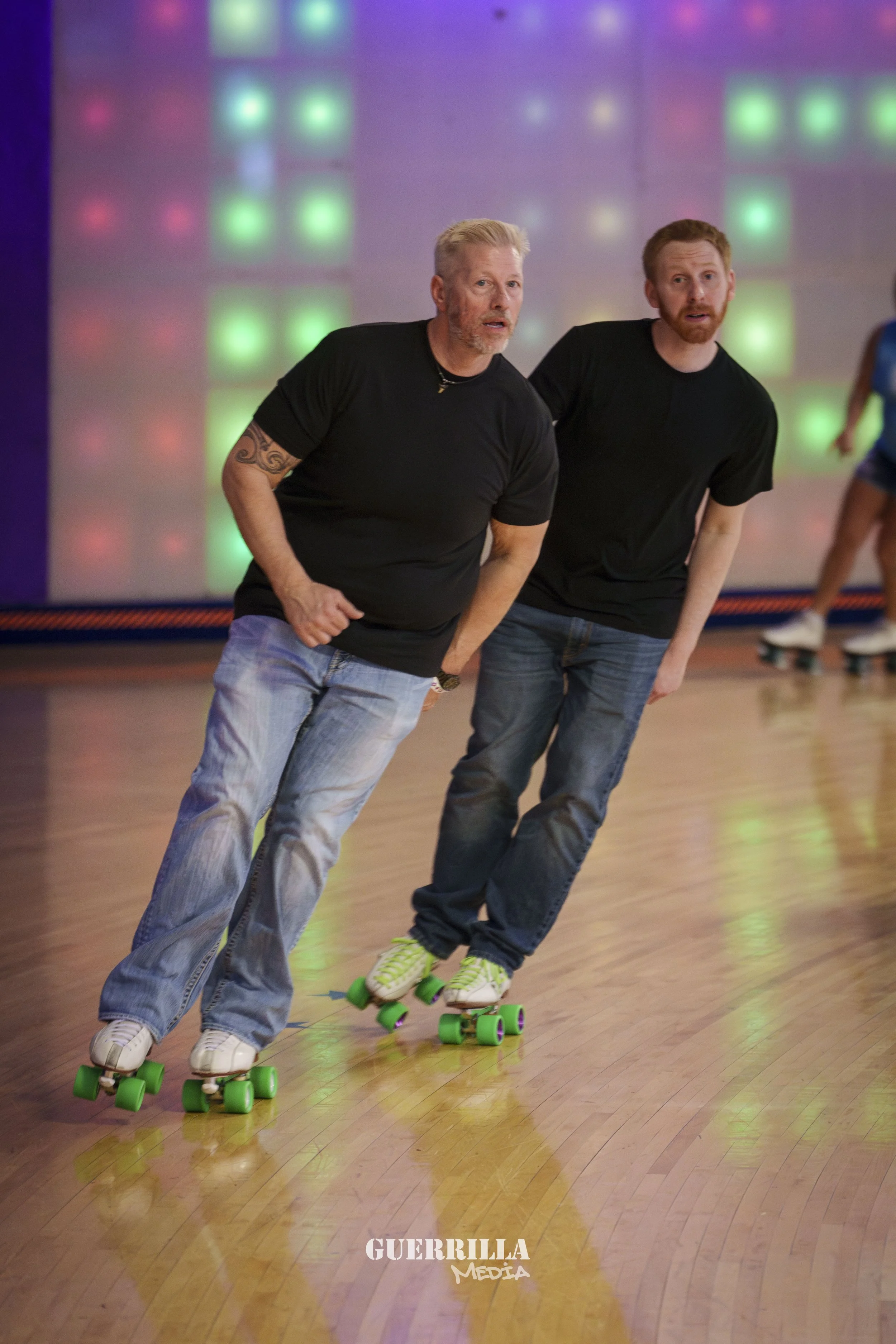 Two men roller skating indoors with a colorful, illuminated wall in the background. The man on the left has gray hair, a beard, and a tattoo sleeve; the man on the right has red hair and a beard. Both are wearing black t-shirts and jeans.