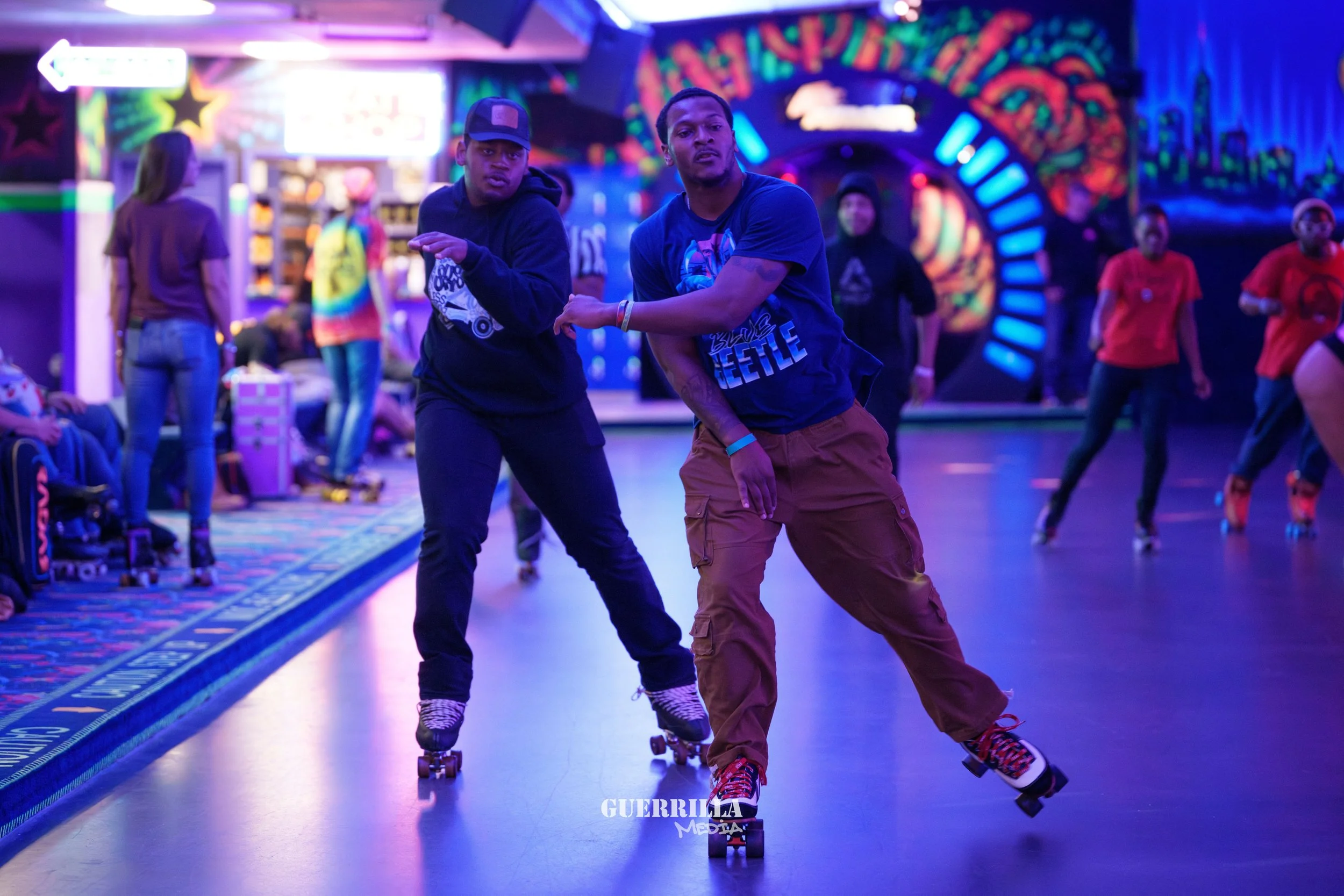 Two young men roller skating together at an indoor roller skating rink with vibrant, colorful neon lights and graffiti-style wall art in the background. Other skaters and spectators are visible.