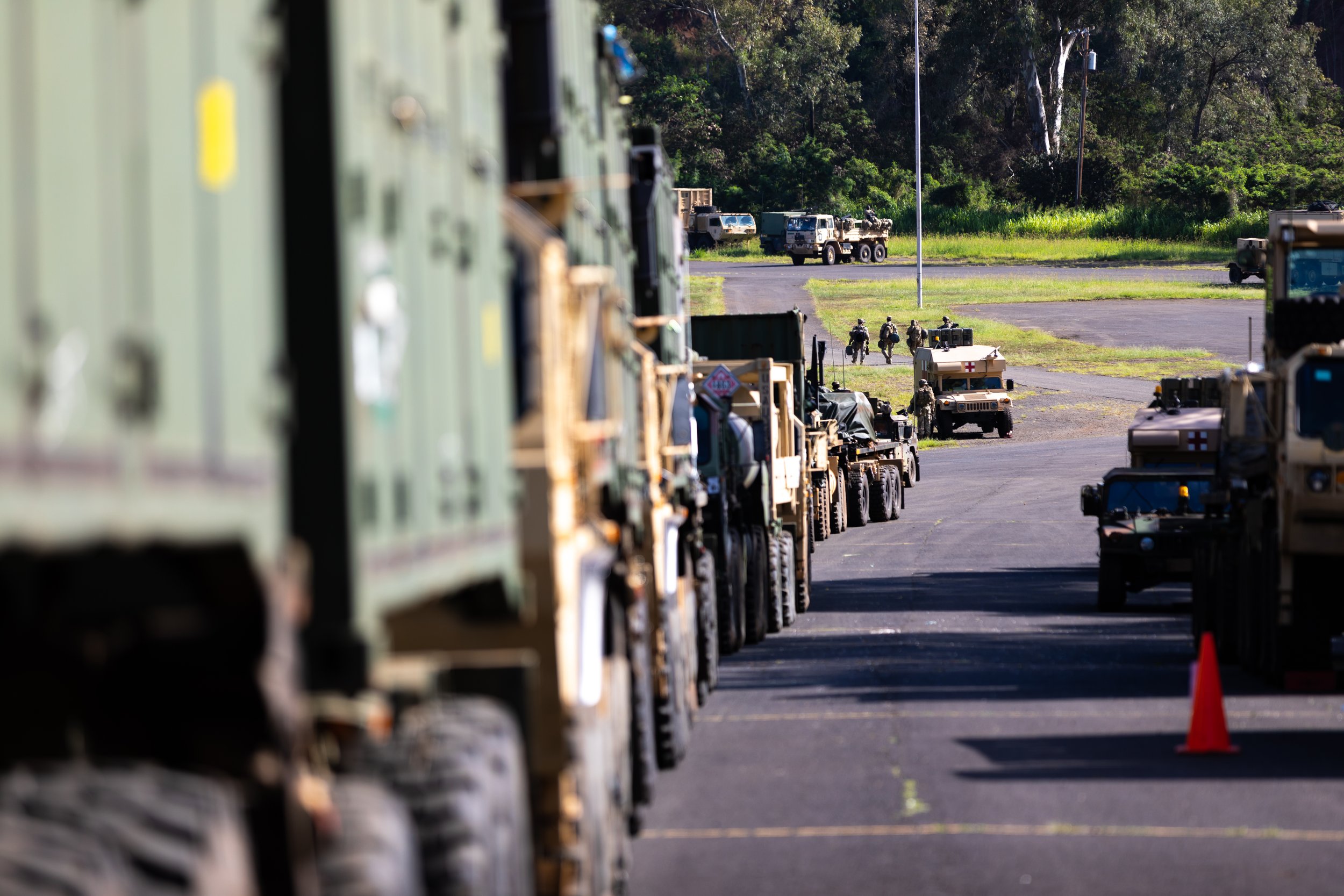 Military vehicles and personnel lined up outdoors on a paved area, with some walking and others near trucks and equipment, surrounded by greenery.