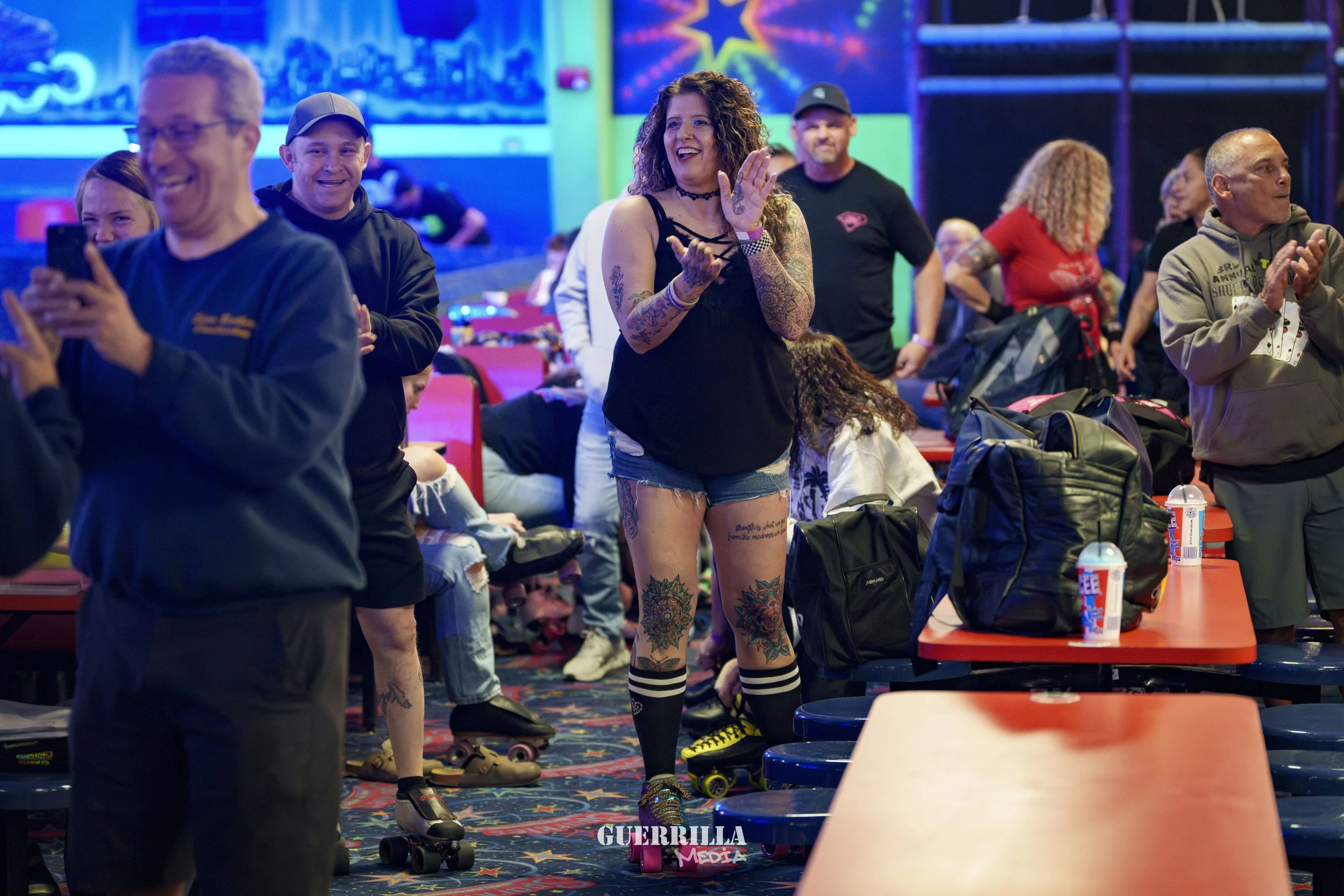 People at an indoor roller skating rink, some standing and some sitting and skating, with vibrant neon lights and a mural of a cityscape in the background.