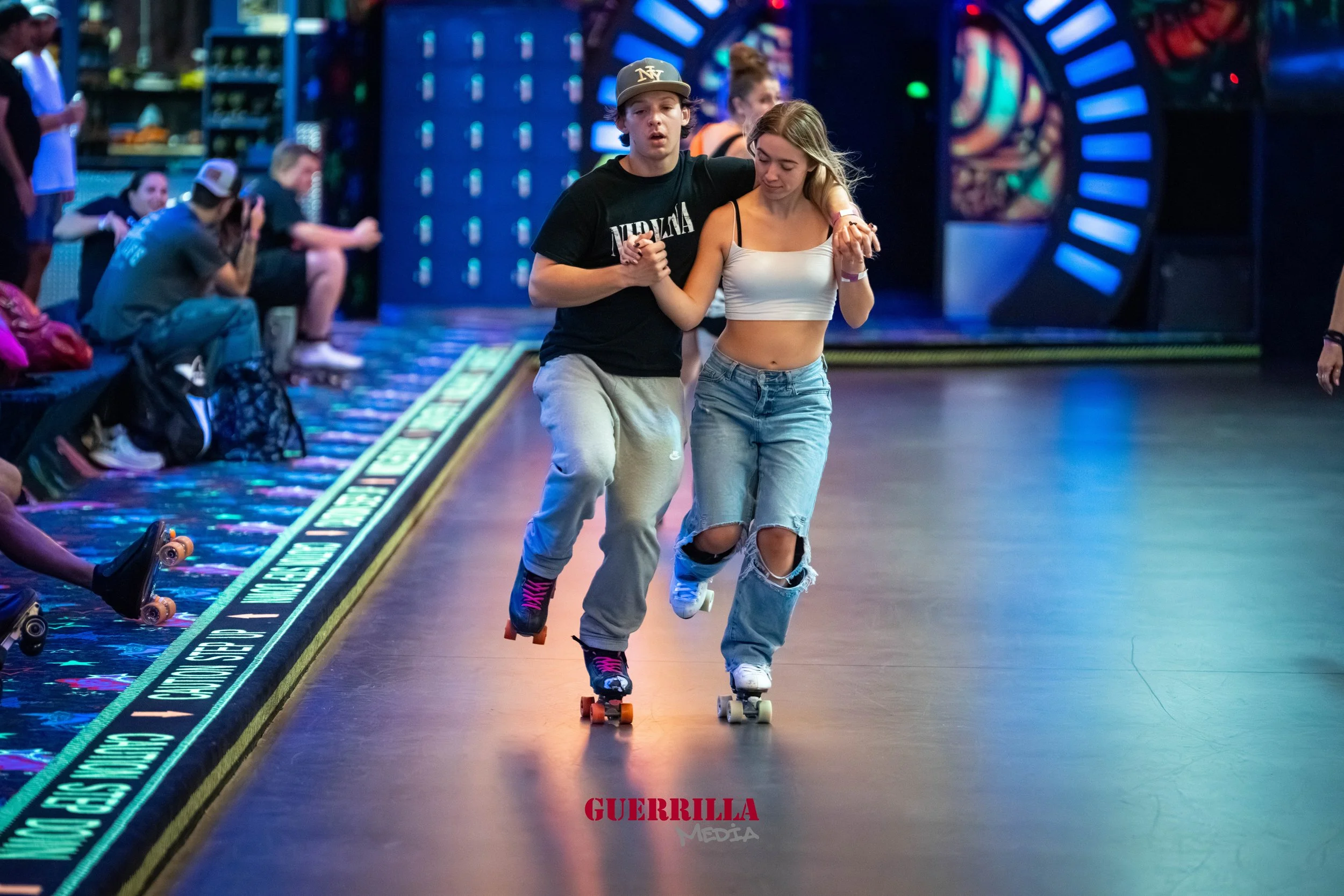 Two young people roller skating together in a skate rink, with other skaters seated along the wall in the background.