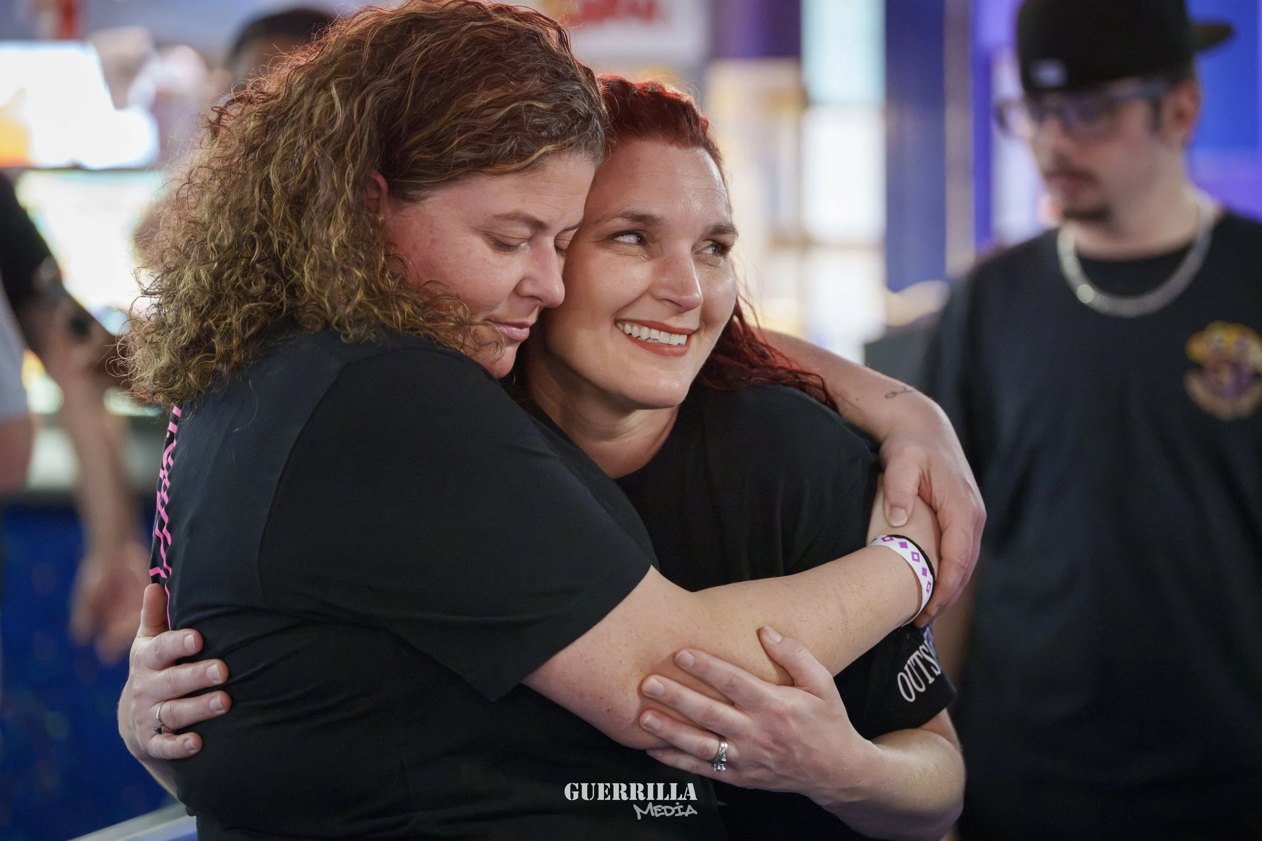 Two women hugging and smiling at an event, with a man in the background looking down.