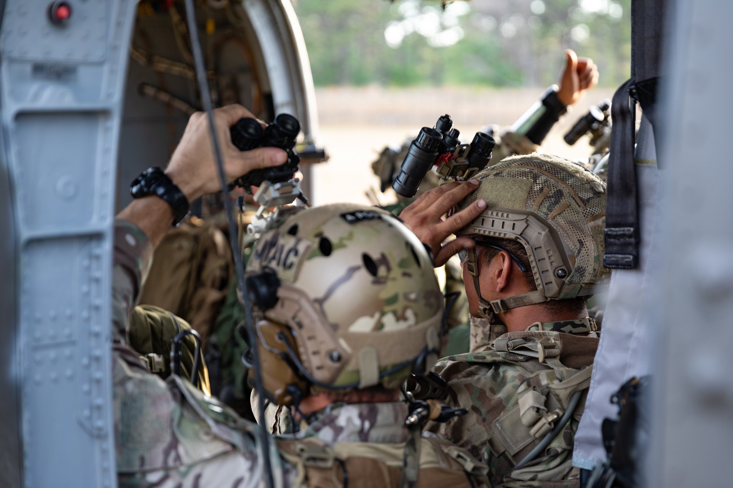 Military personnel in camouflage uniforms and helmets inside a helicopter, using binoculars and communication devices, with one adjusting equipment and another looking through binoculars.