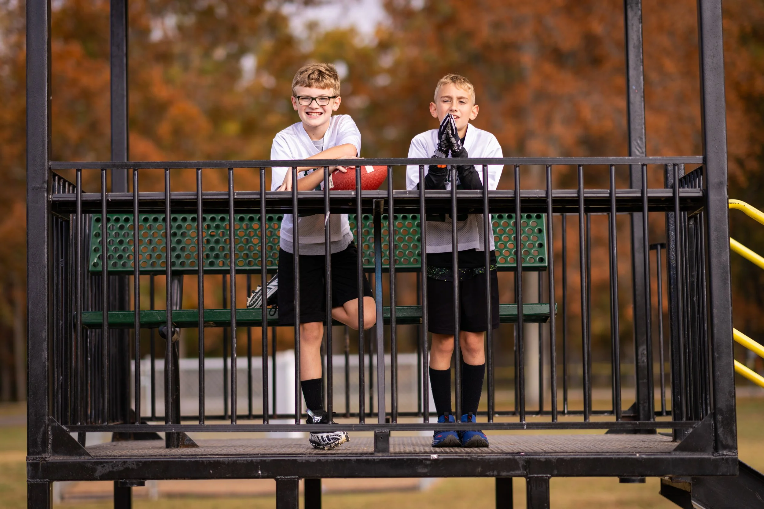 Two boys in football uniforms standing on a playground platform behind black metal railing, one holding a football and the other with hands in a praying pose, with autumn trees in the background.