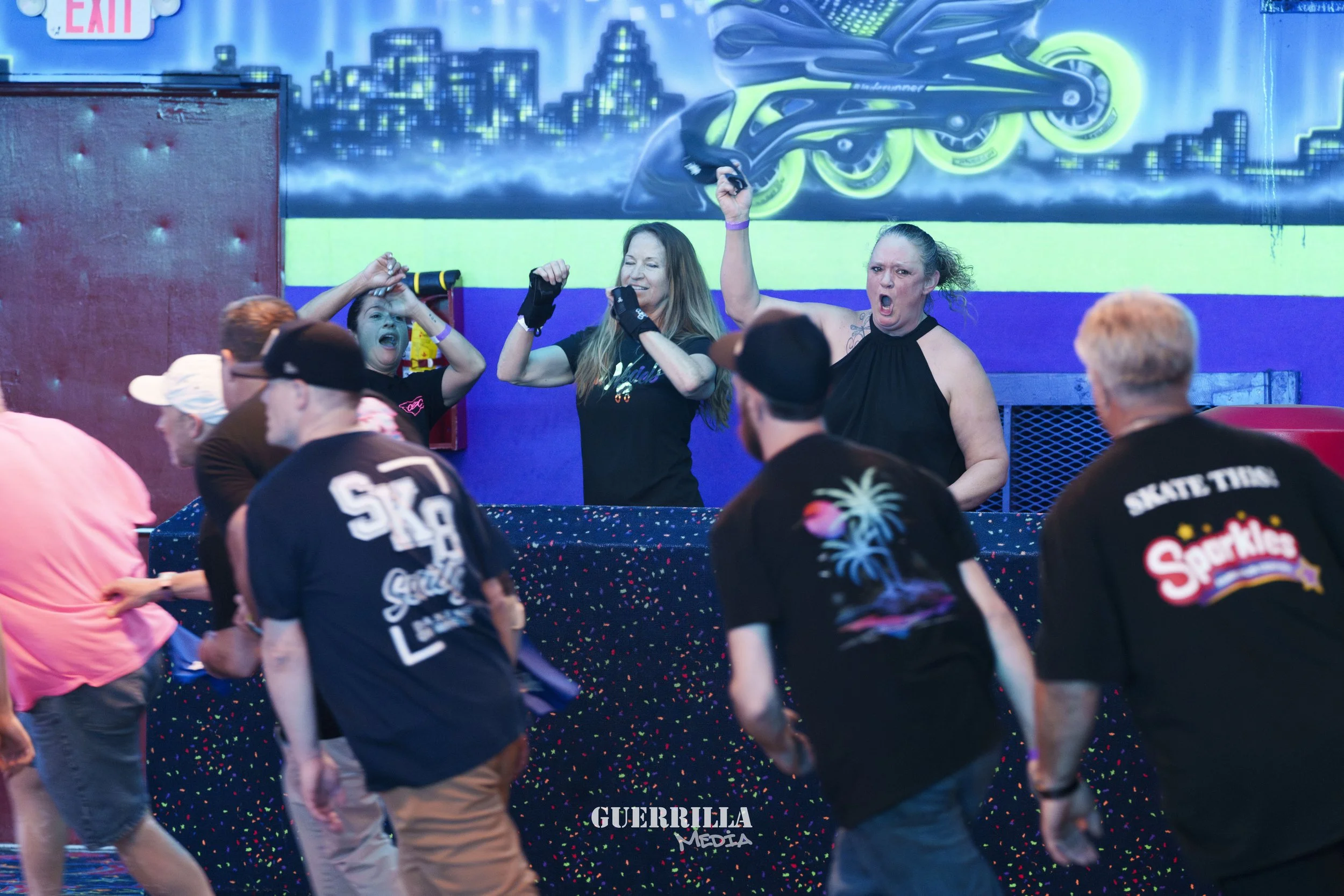 People skating indoors at a roller skating rink, with some dancing or celebrating behind a counter with a colorful backdrop of city skyline and neon rollerblades.