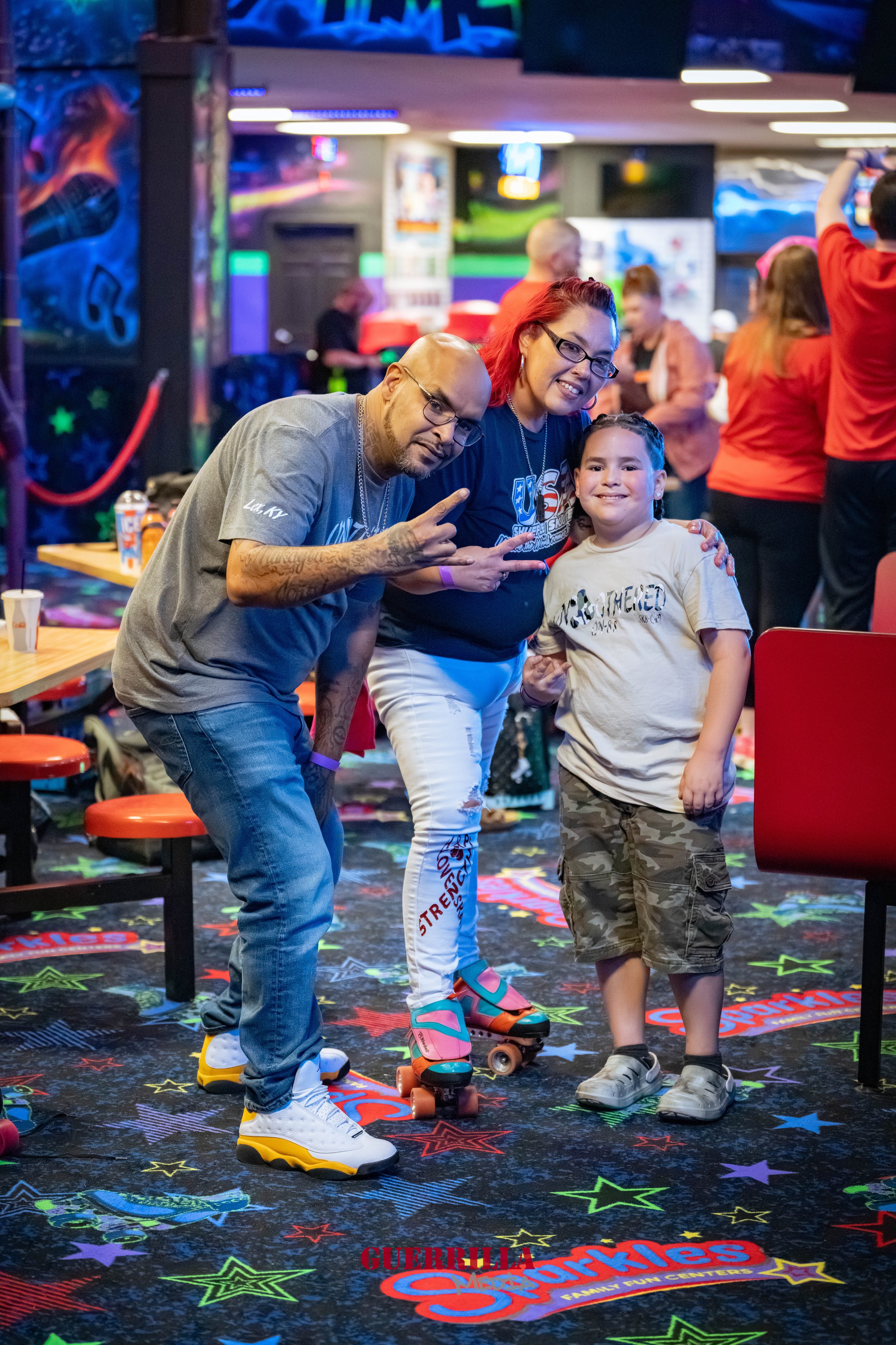 A group of three people, a man, a woman, and a young boy, at an arcade or entertainment center. The boy is on roller skates, smiling, with the man and woman posing beside him, all making peace signs. The background contains colorful lights, arcade de