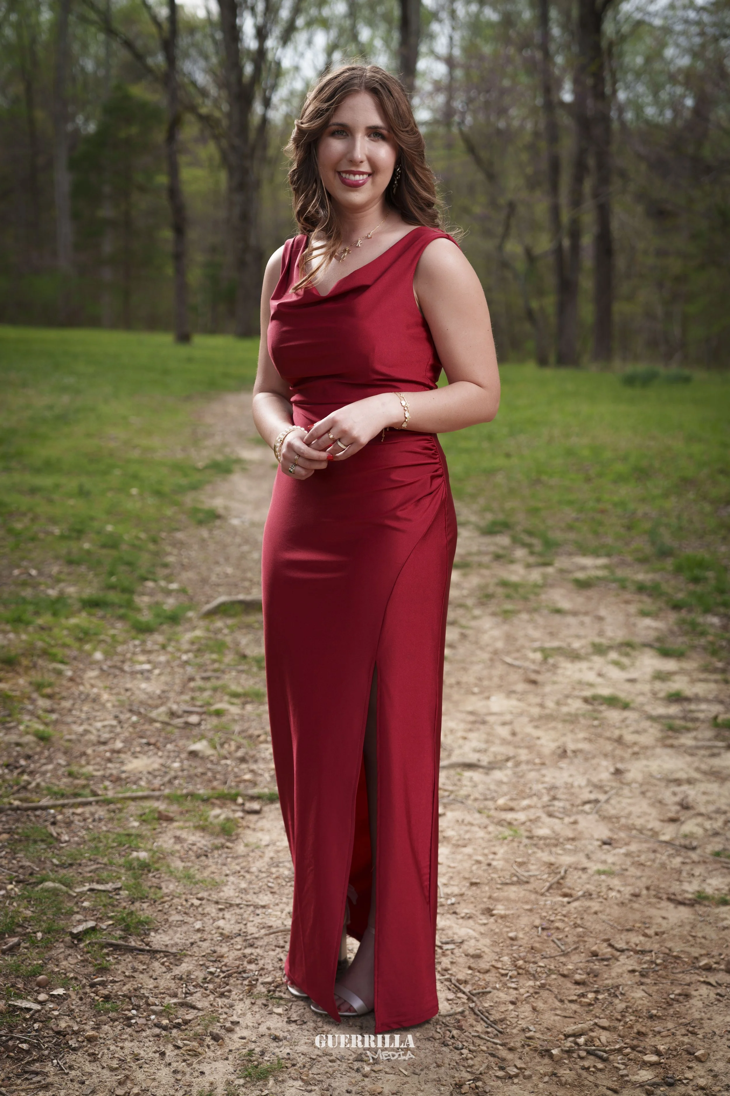 A young woman in a sleeveless red gown standing outdoors on a dirt path in a wooded area, smiling at the camera.