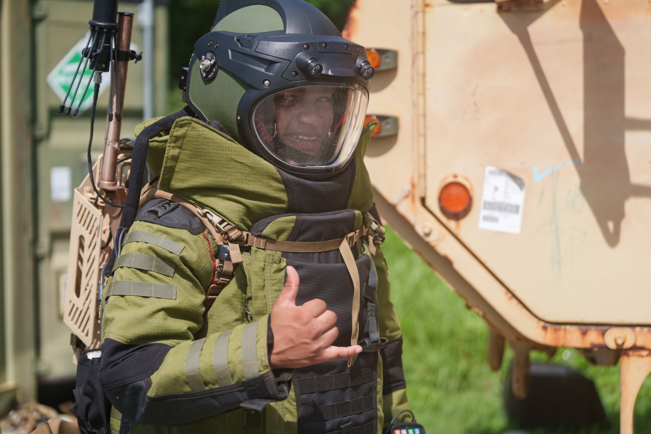 A person in military attire giving a thumbs-up, wearing a helmet with a face shield and a green tactical jacket, standing outdoors near a large metal vehicle or equipment.