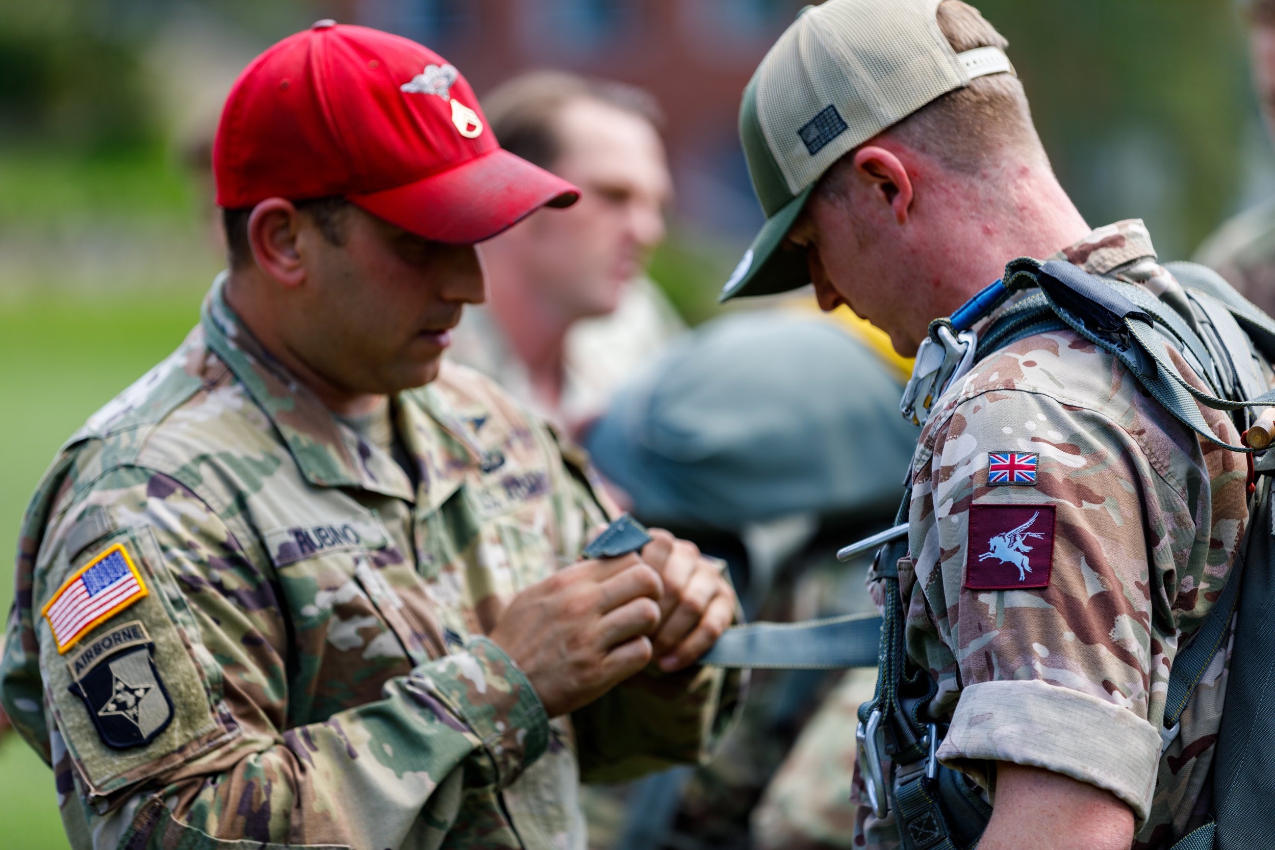 Two soldiers in camouflage uniforms with patches, one with a red cap and the other with a beige cap, standing outdoors with a green background, preparing equipment