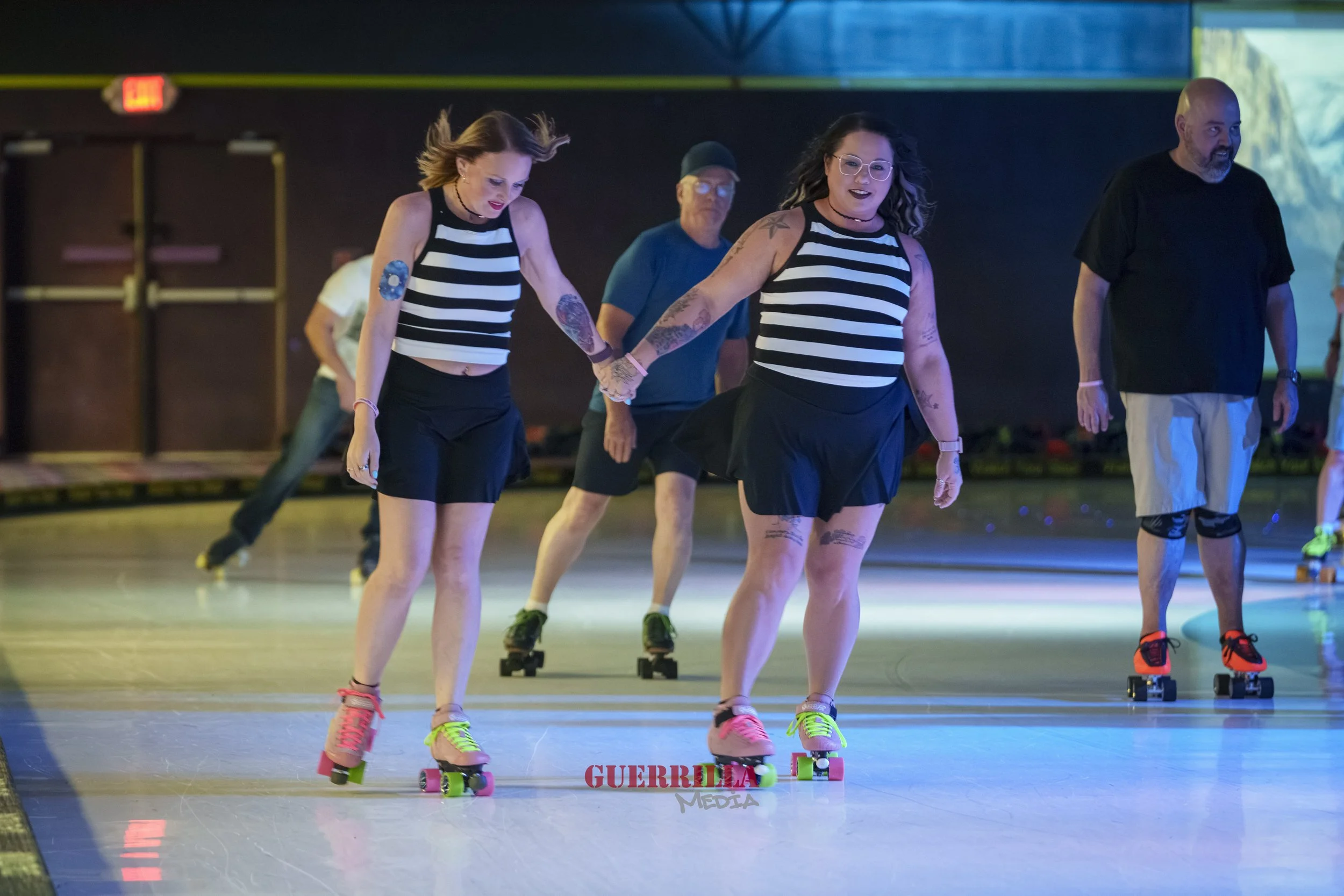 Two women roller skating while holding hands at an indoor roller rink with other skaters in the background.
