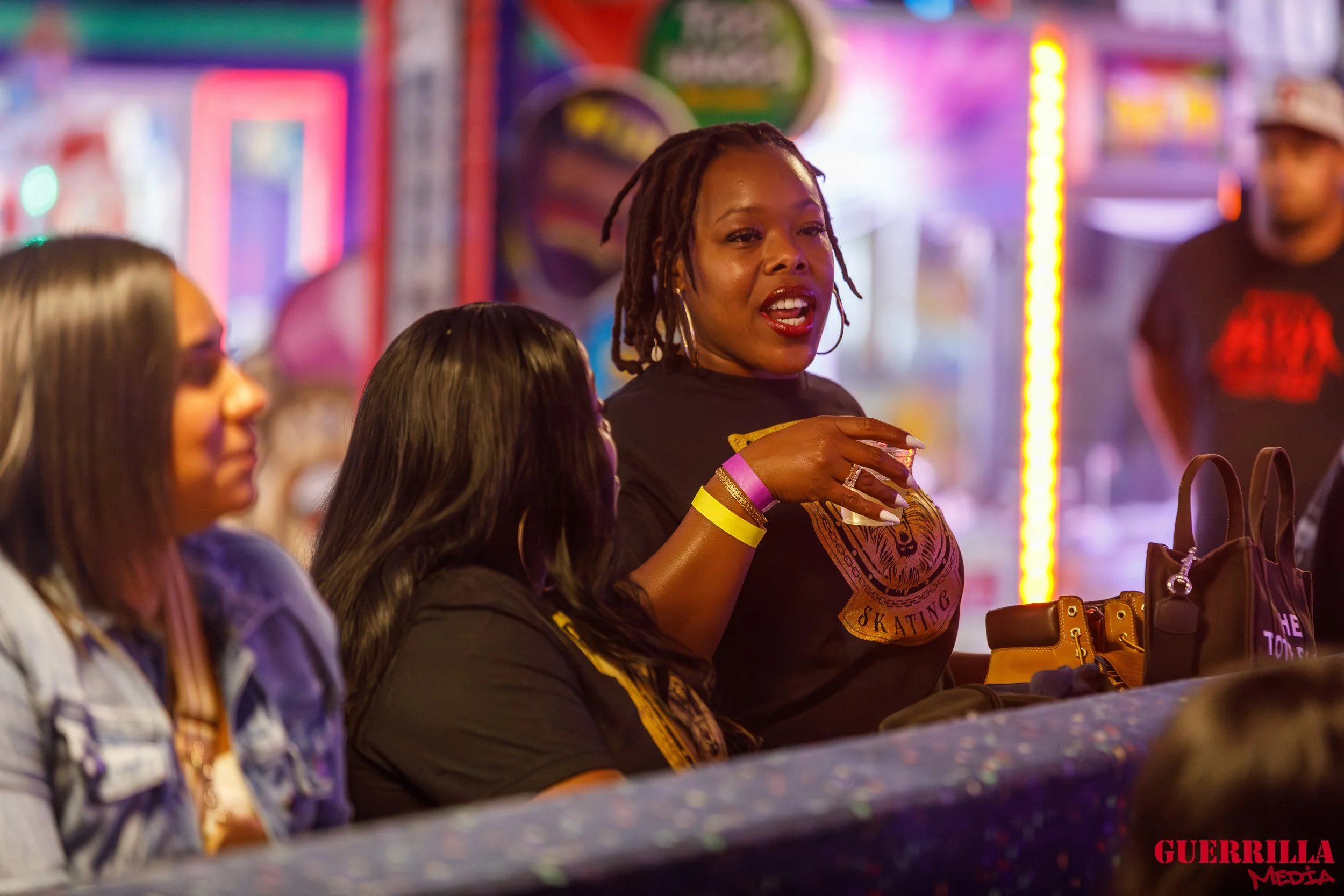 People sitting at a booth or bar in a vibrant, neon-lit setting at night, with one woman speaking and gesturing, colorful wristbands visible, bags on the table, and a blurred person in the background.