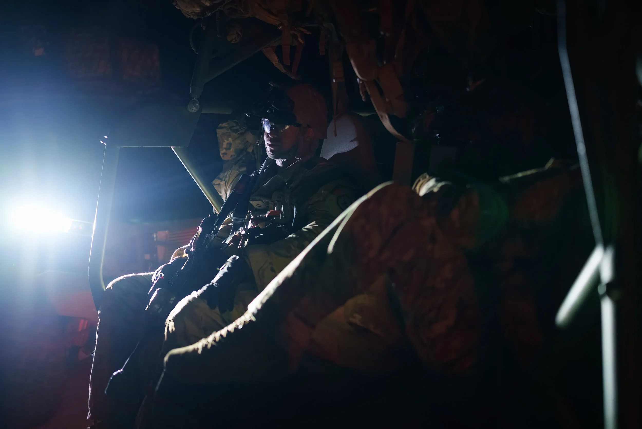 A soldier in camouflage gear sitting inside a military aircraft cockpit, holding a rifle, illuminated by a bright light from outside.