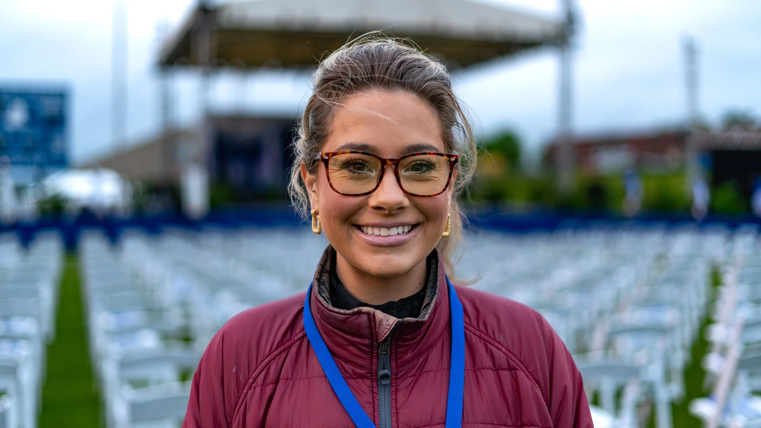 Smiling woman with glasses and earrings wearing a maroon jacket at an outdoor event with chairs and a stage in the background.
