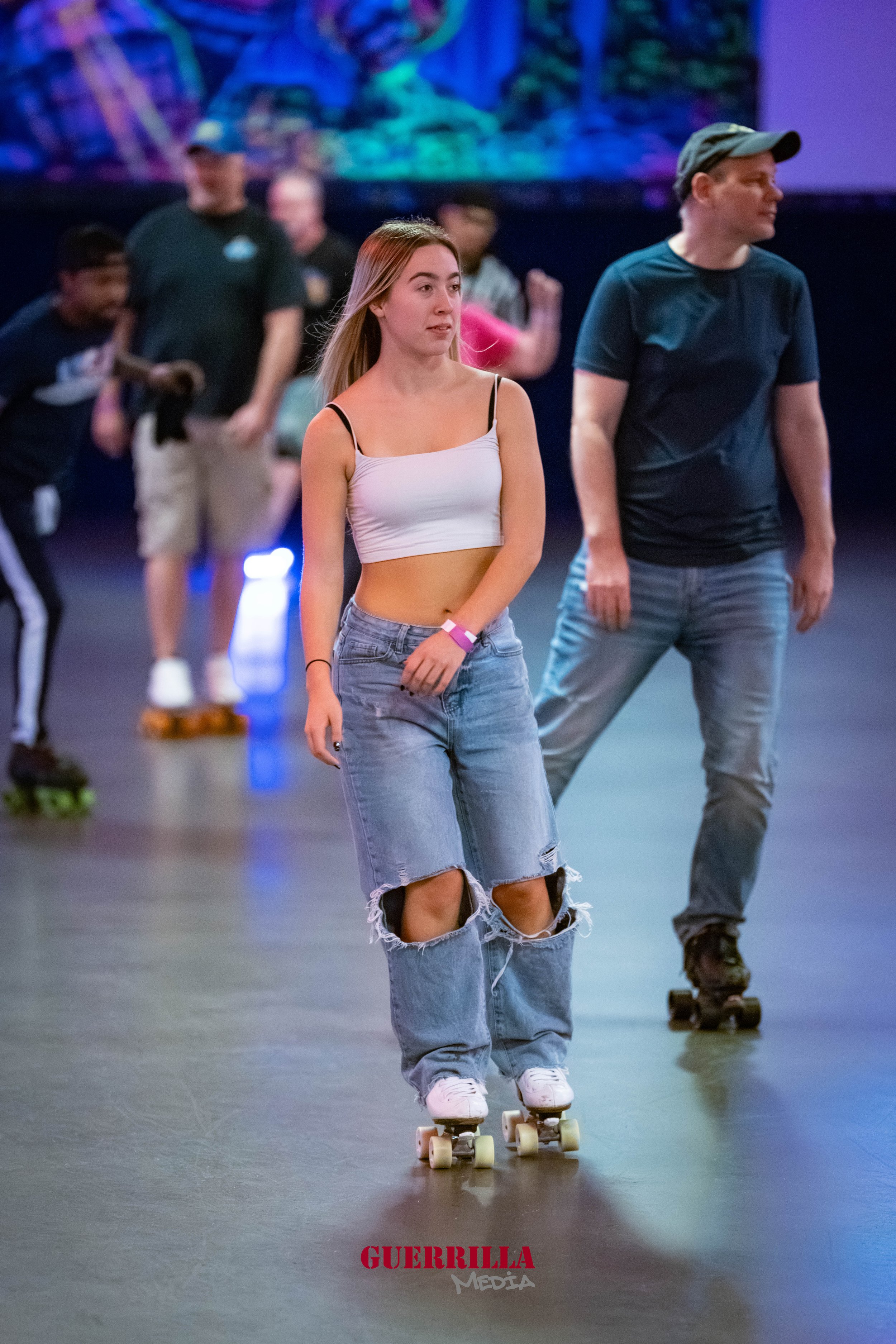 Young woman skateboarding indoors, wearing ripped jeans and a white crop top, with several people in the background also skateboarding and wearing casual clothing.