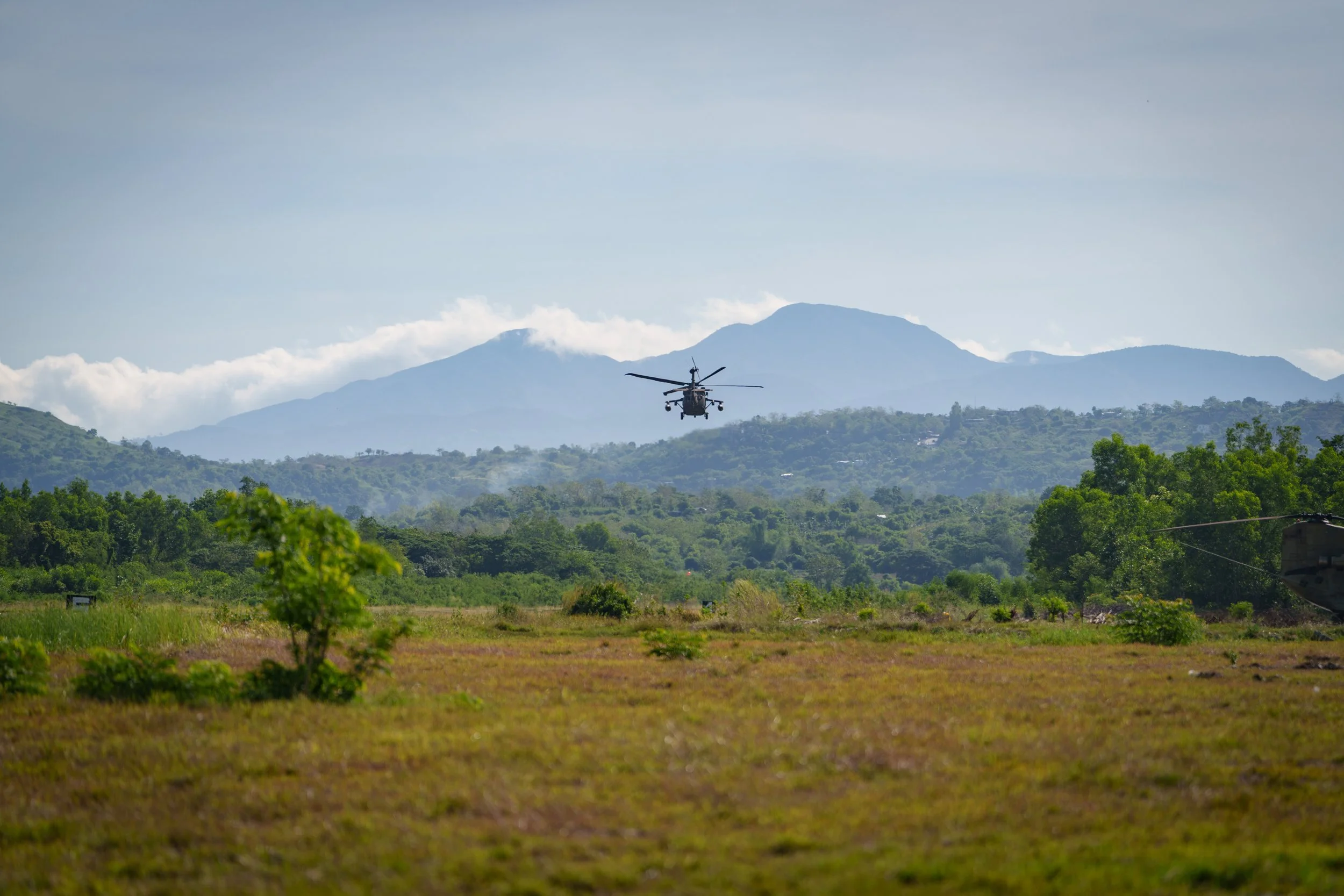 A helicopter flying over a grassy field with trees and mountains in the background.