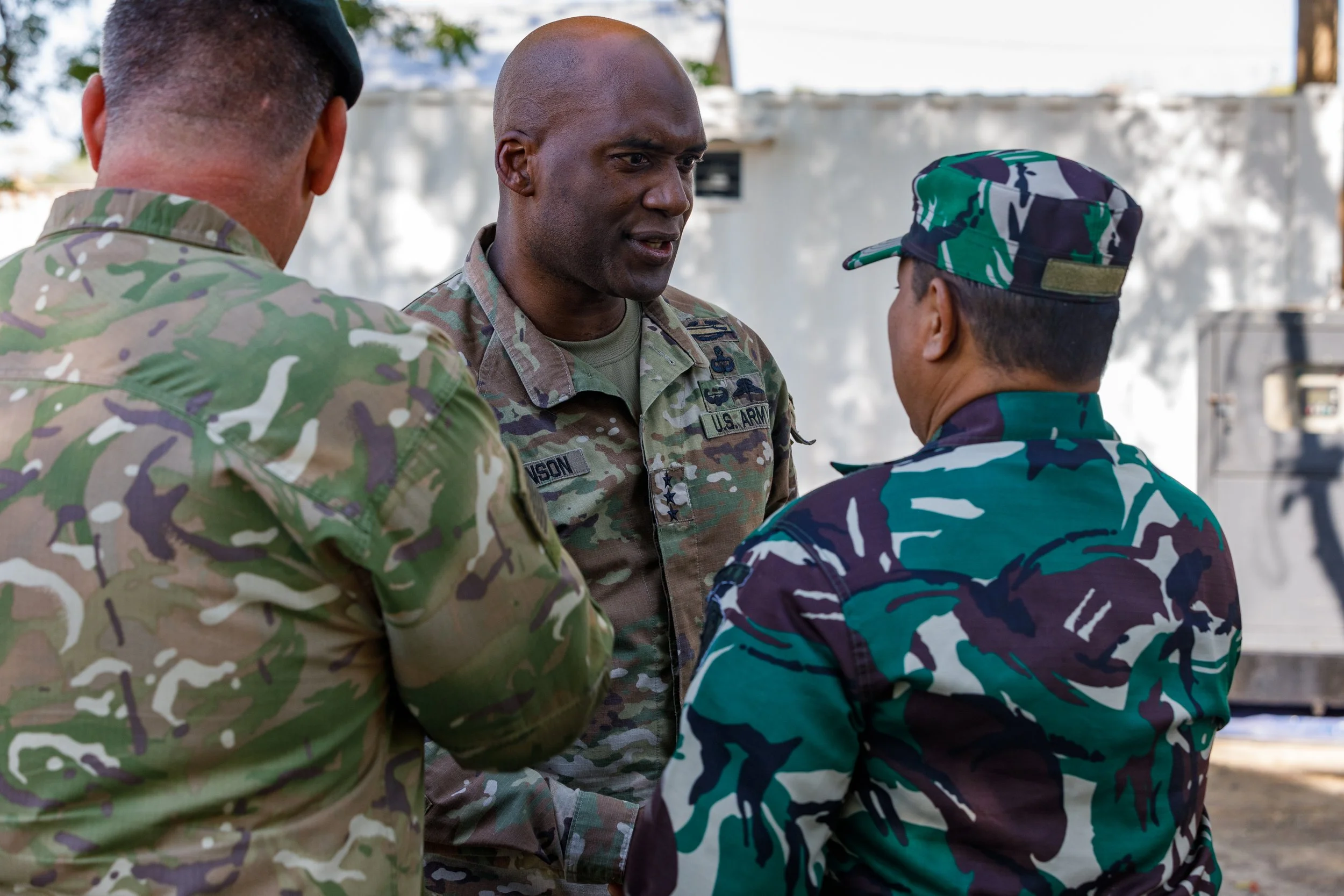 Three military personnel in camouflage uniforms having a conversation outdoors.