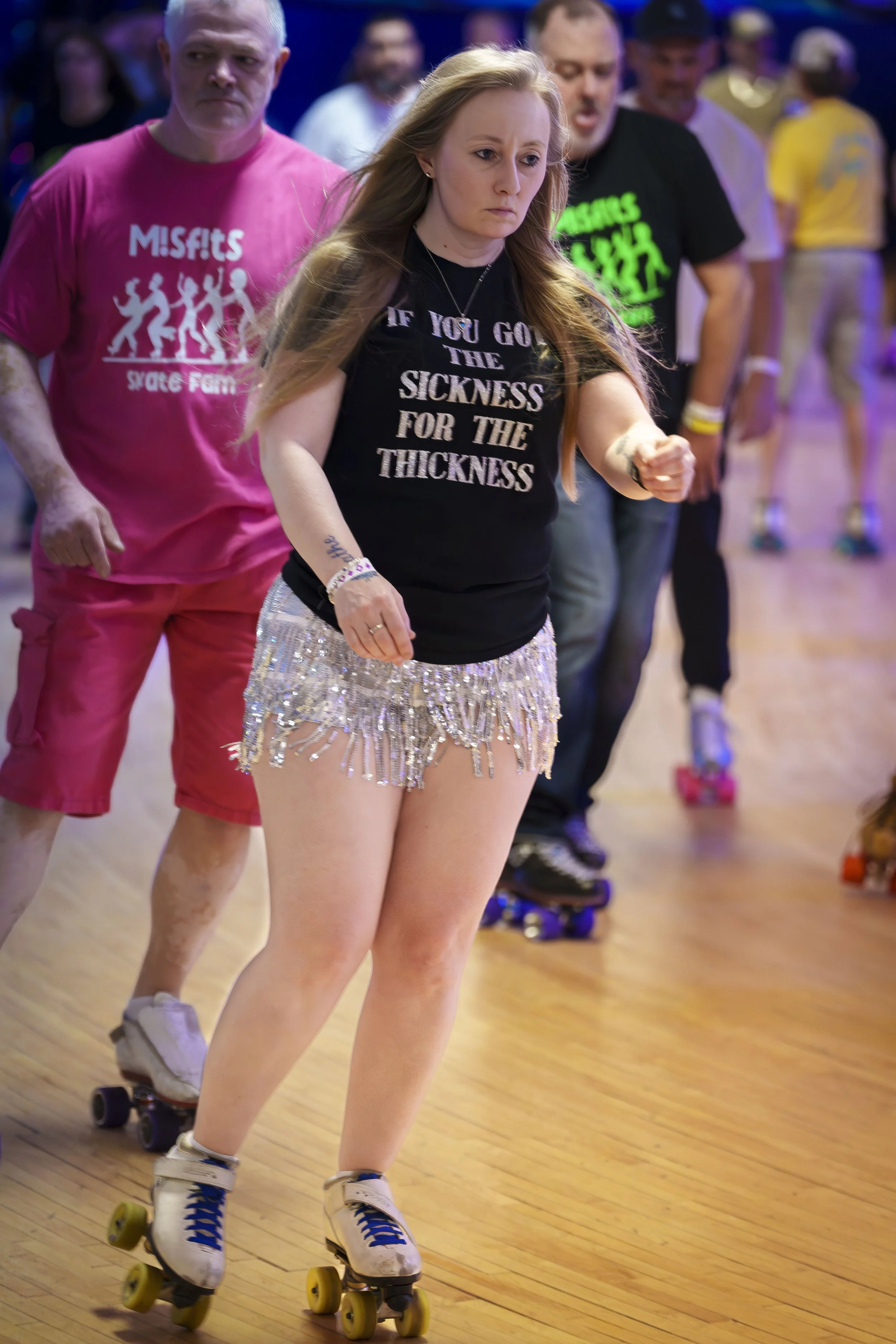 A woman rollerskating in a line with others in the background at an indoor skating rink, wearing a black T-shirt with white text and shiny short shorts.