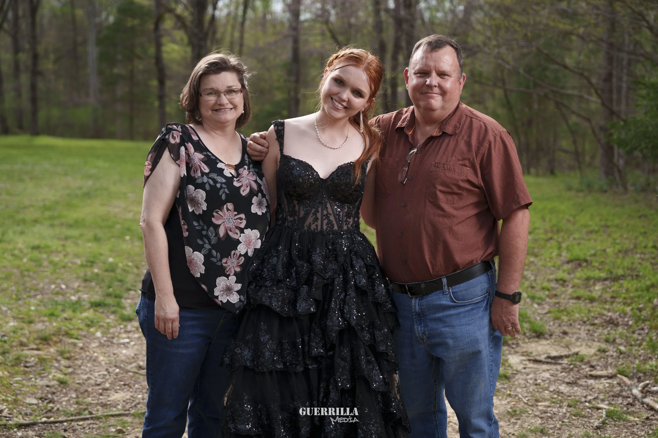 A young woman in a black, lace, and sequin gown stands between an older woman with glasses wearing a floral blouse and a man in a maroon shirt and jeans, outdoors with trees and grass in the background.