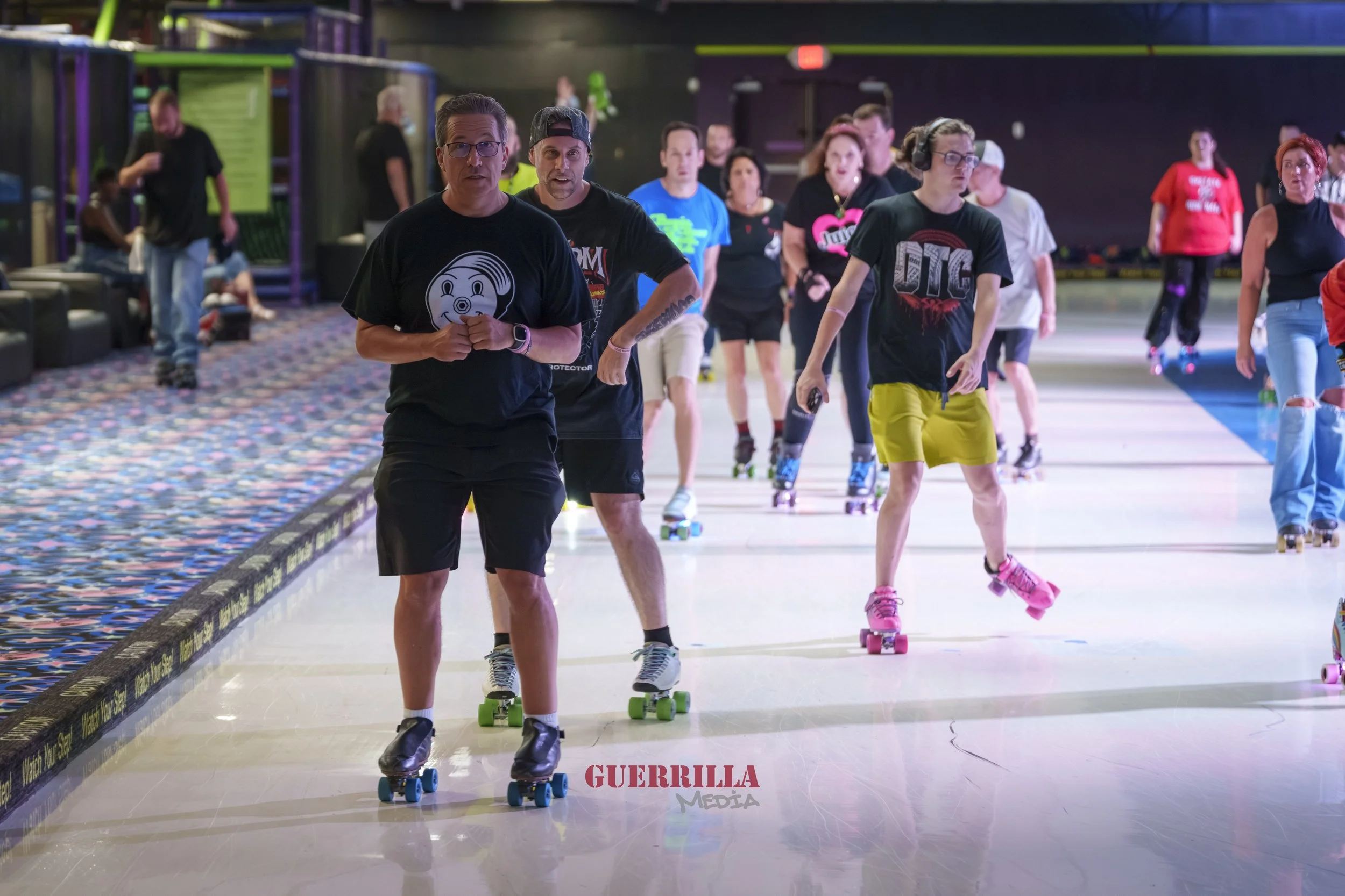 People roller skating in an indoor rink, some wearing casual workout clothes, with a colorful patterned carpet on the side and dark walls in the background.