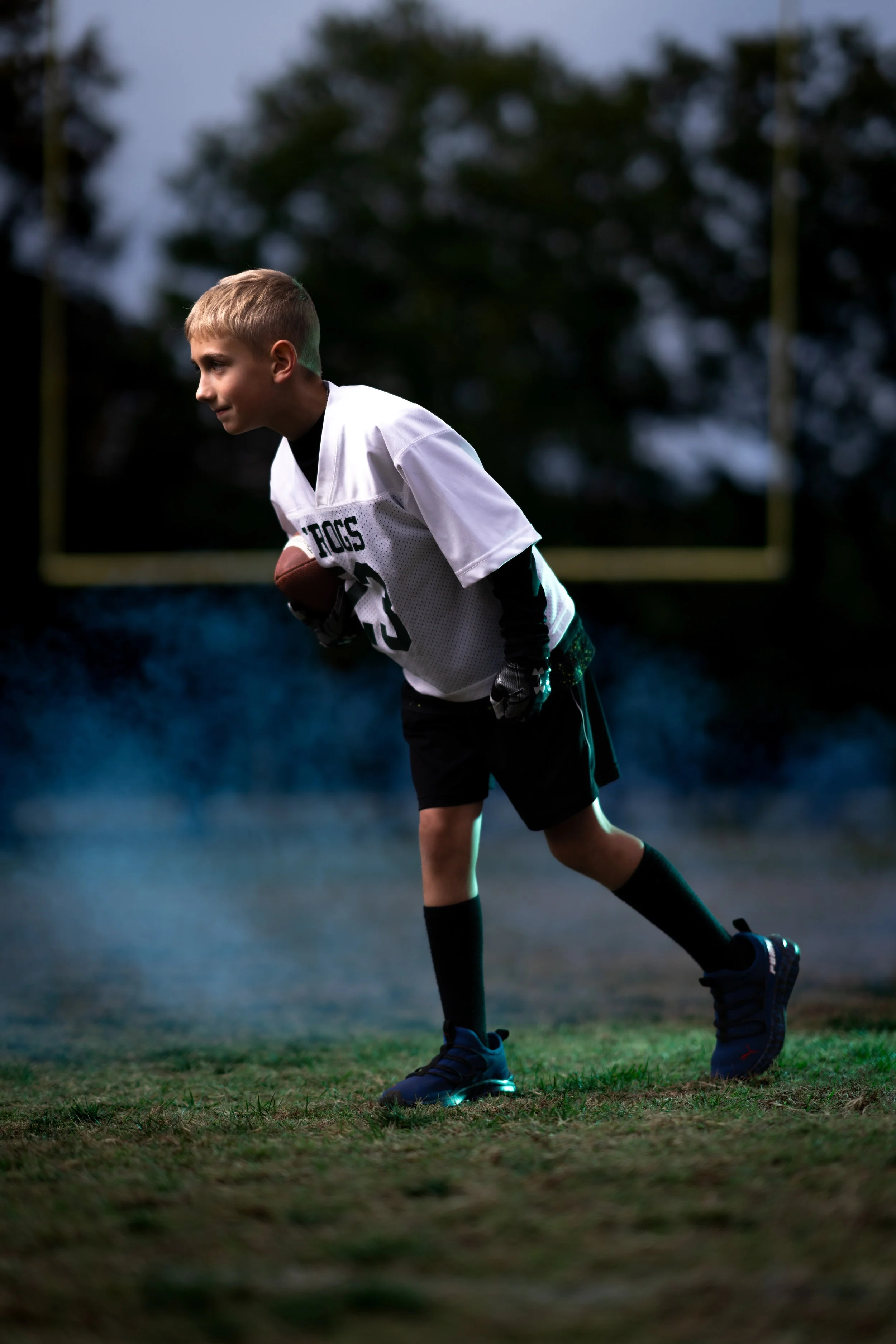 Young boy in football uniform holding a football on a field at dusk.