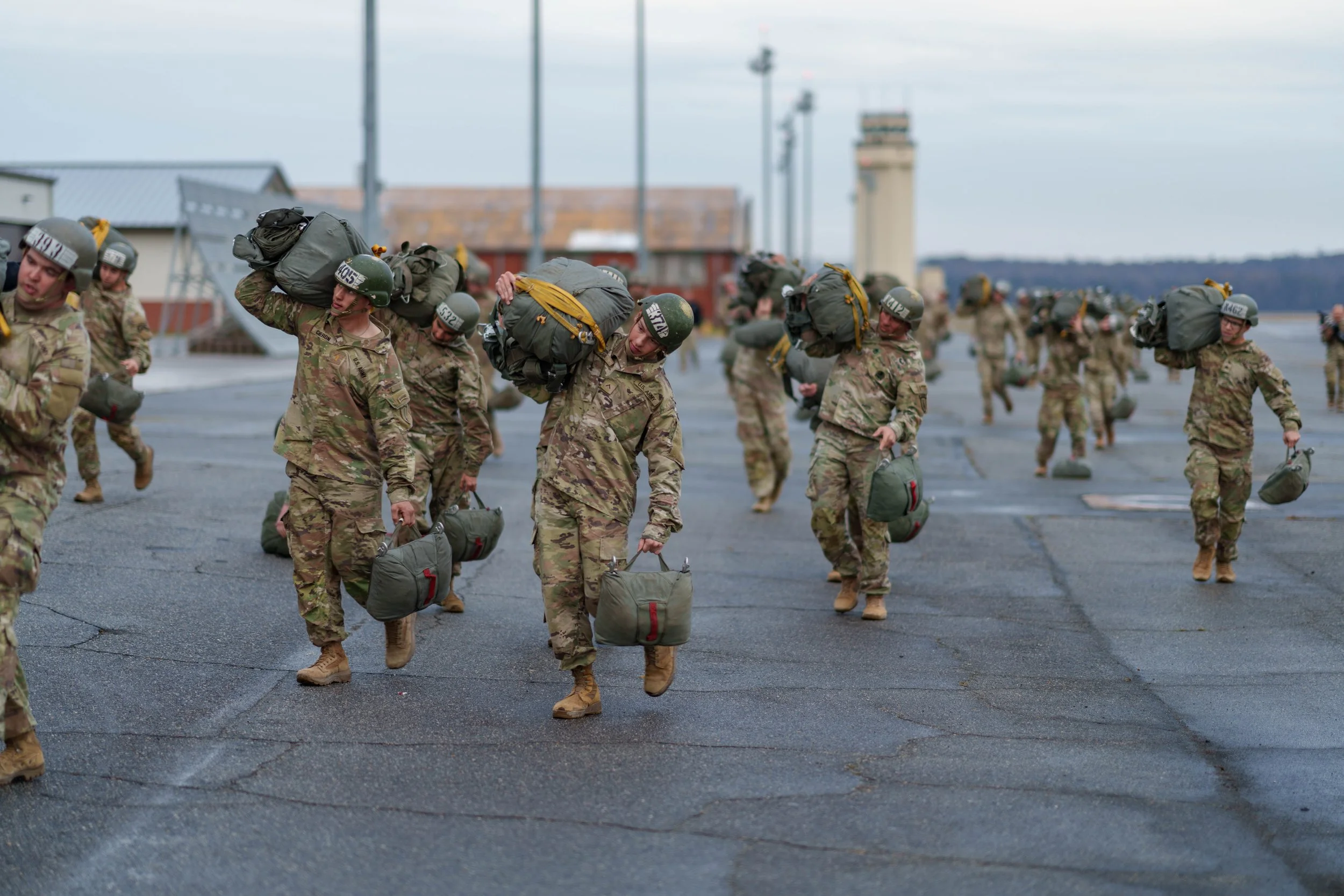 Military personnel walking on an airstrip while carrying gear and helmets.