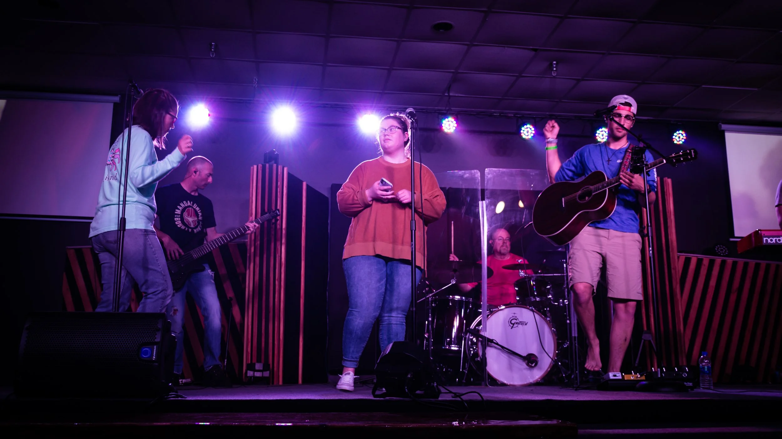 A band performing on stage under purple stage lights. The band includes a female vocalist, a bassist, a drummer behind a clear drum shield, a guitarist, and a keyboardist. The stage has striped wood panels and microphones.