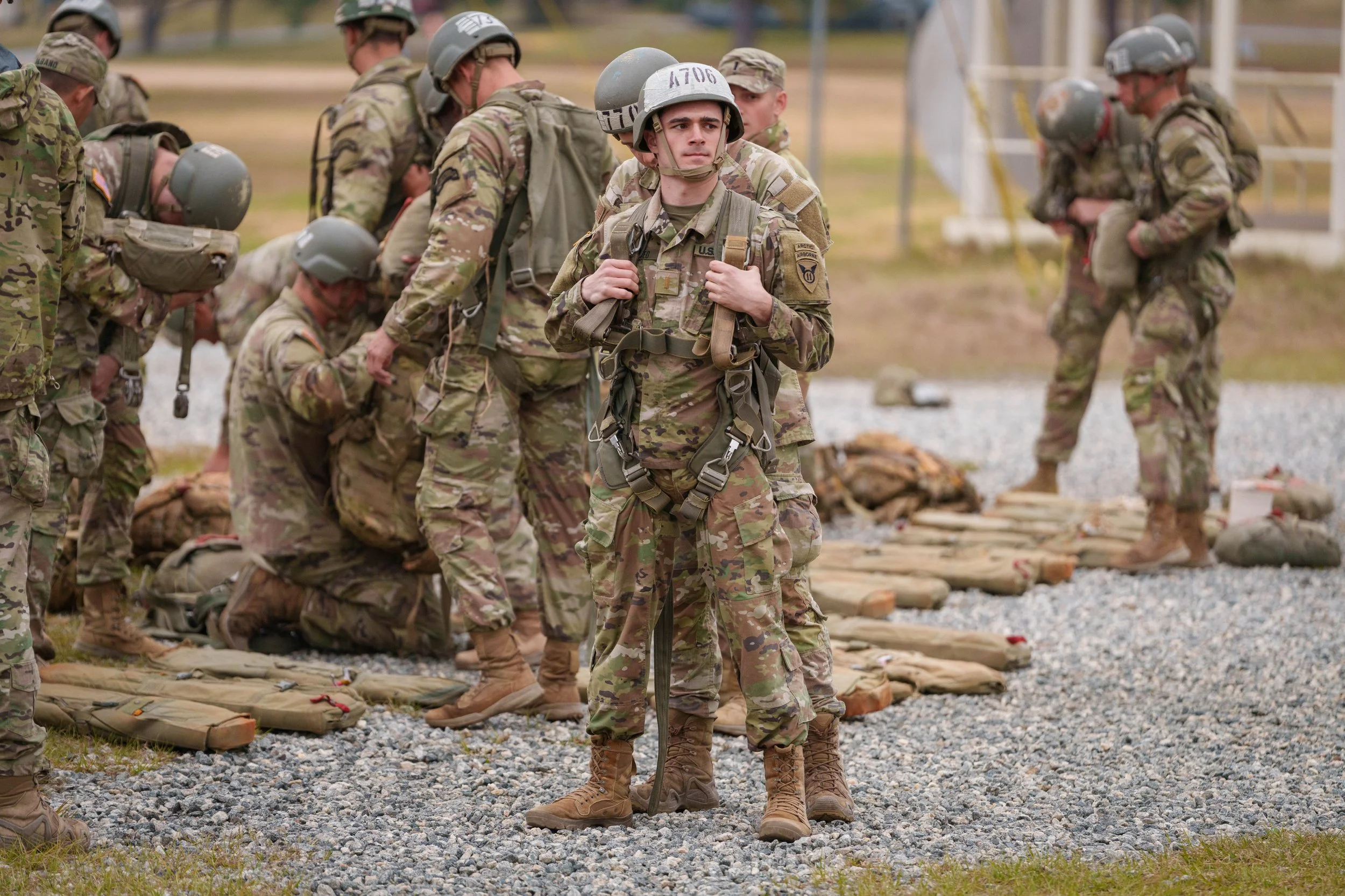 Group of soldiers in camouflage uniforms and helmets standing and kneeling on a gravel surface, with one soldier standing in the foreground holding a backpack, and others praying or preparing nearby.
