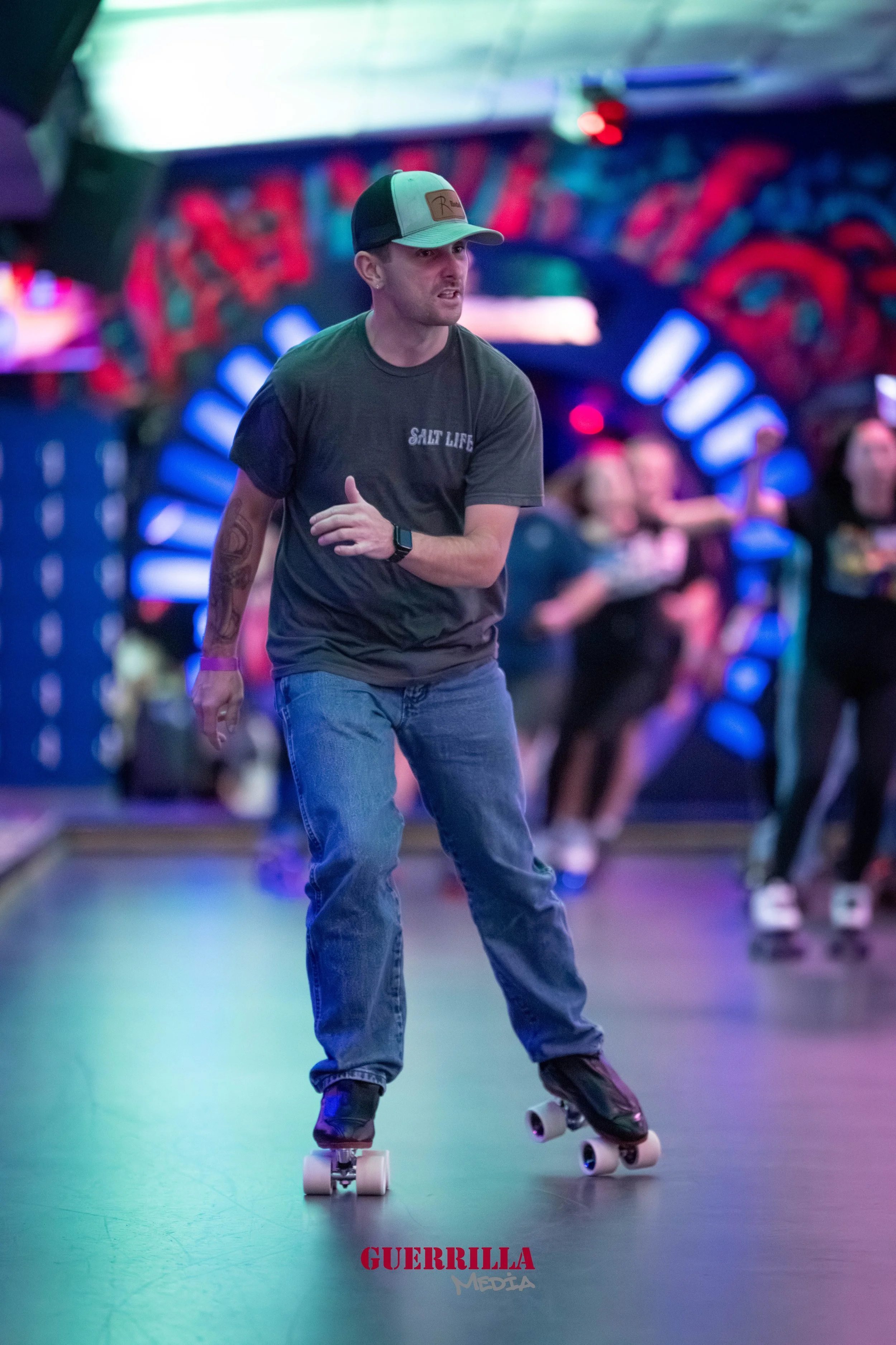 A man skateboarding indoors at a roller rink with colorful graffiti-style art and other skaters in the background.