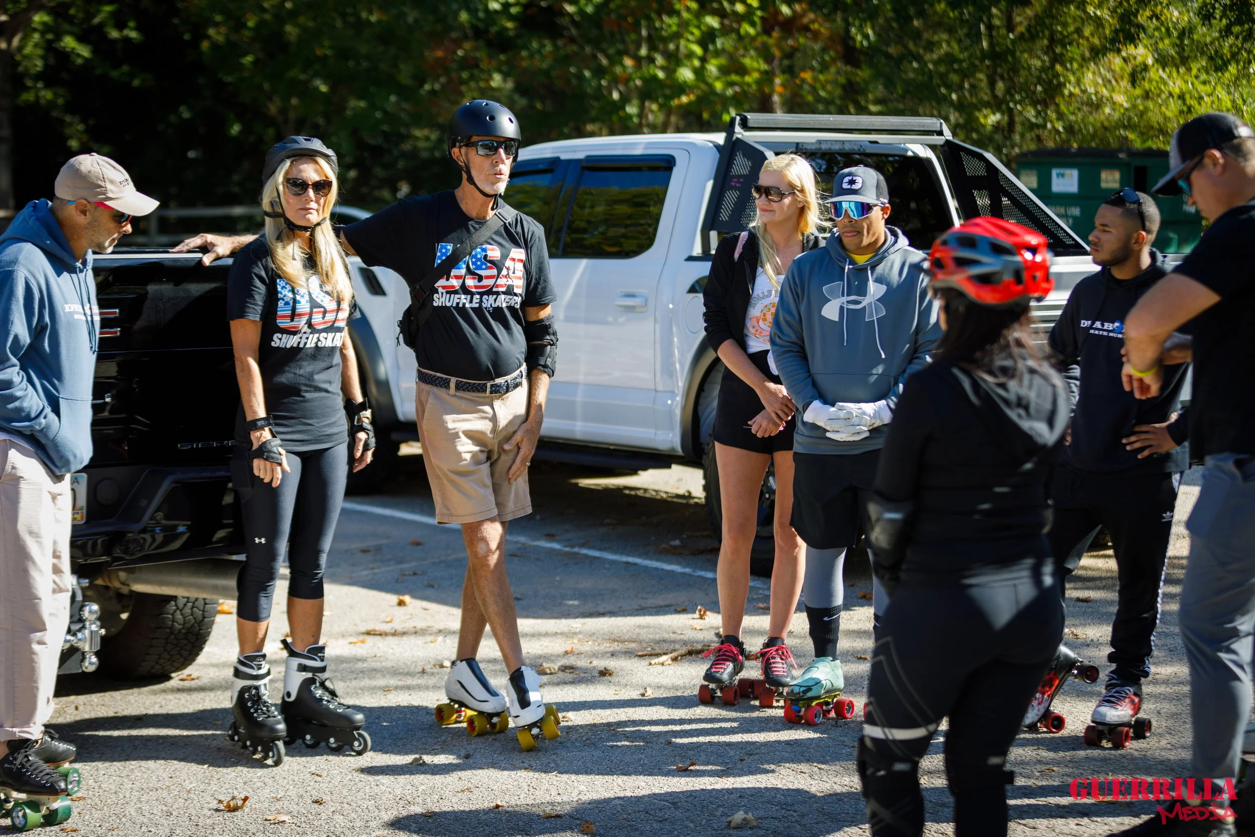 A group of people standing outdoors with skateboards, some wearing helmets and protective gear, engaging in a skater gathering or lesson.