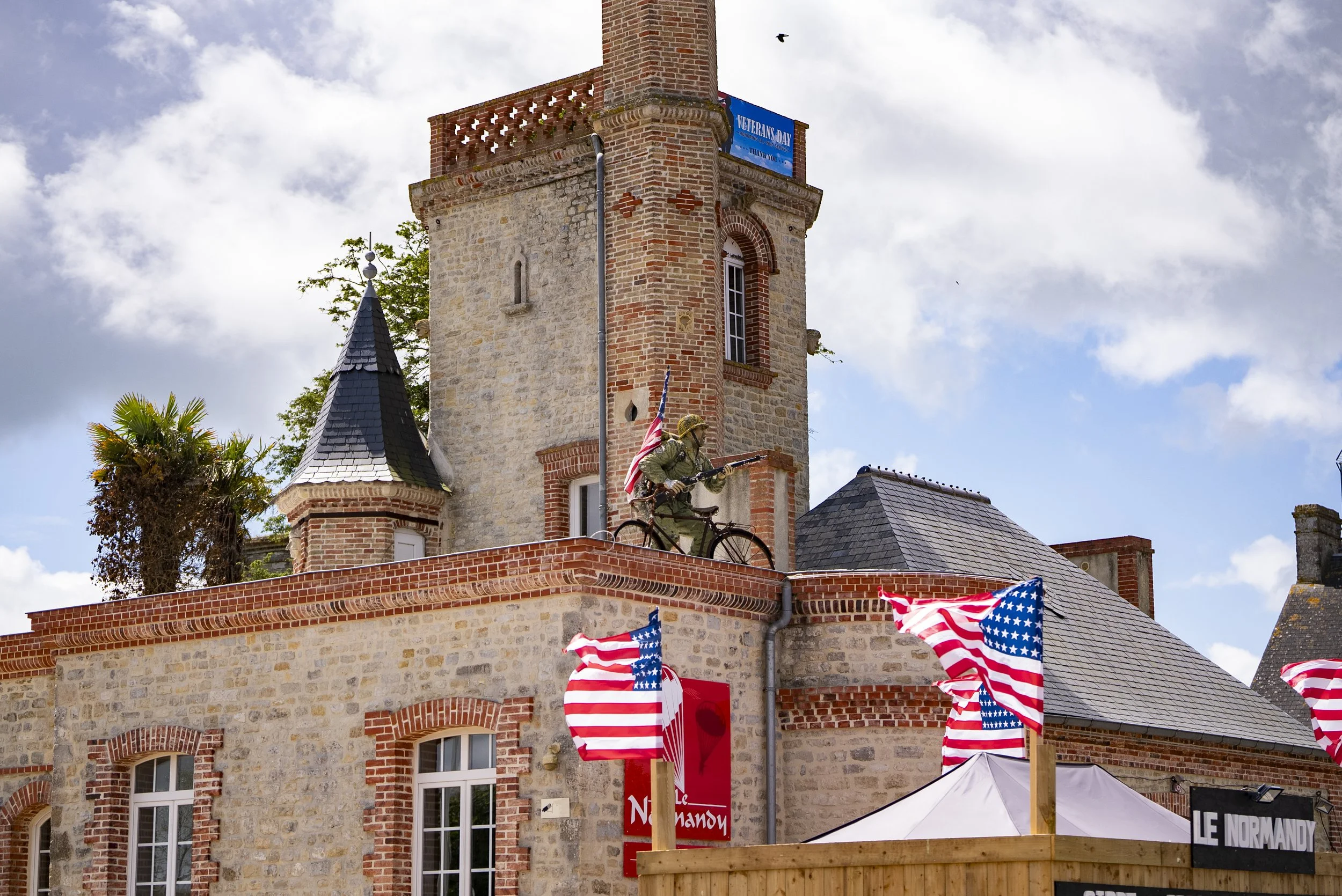 A brick building with a turret and a soldier mannequin holding a rifle on a bicycle, with American flags and a Veterans Day banner.