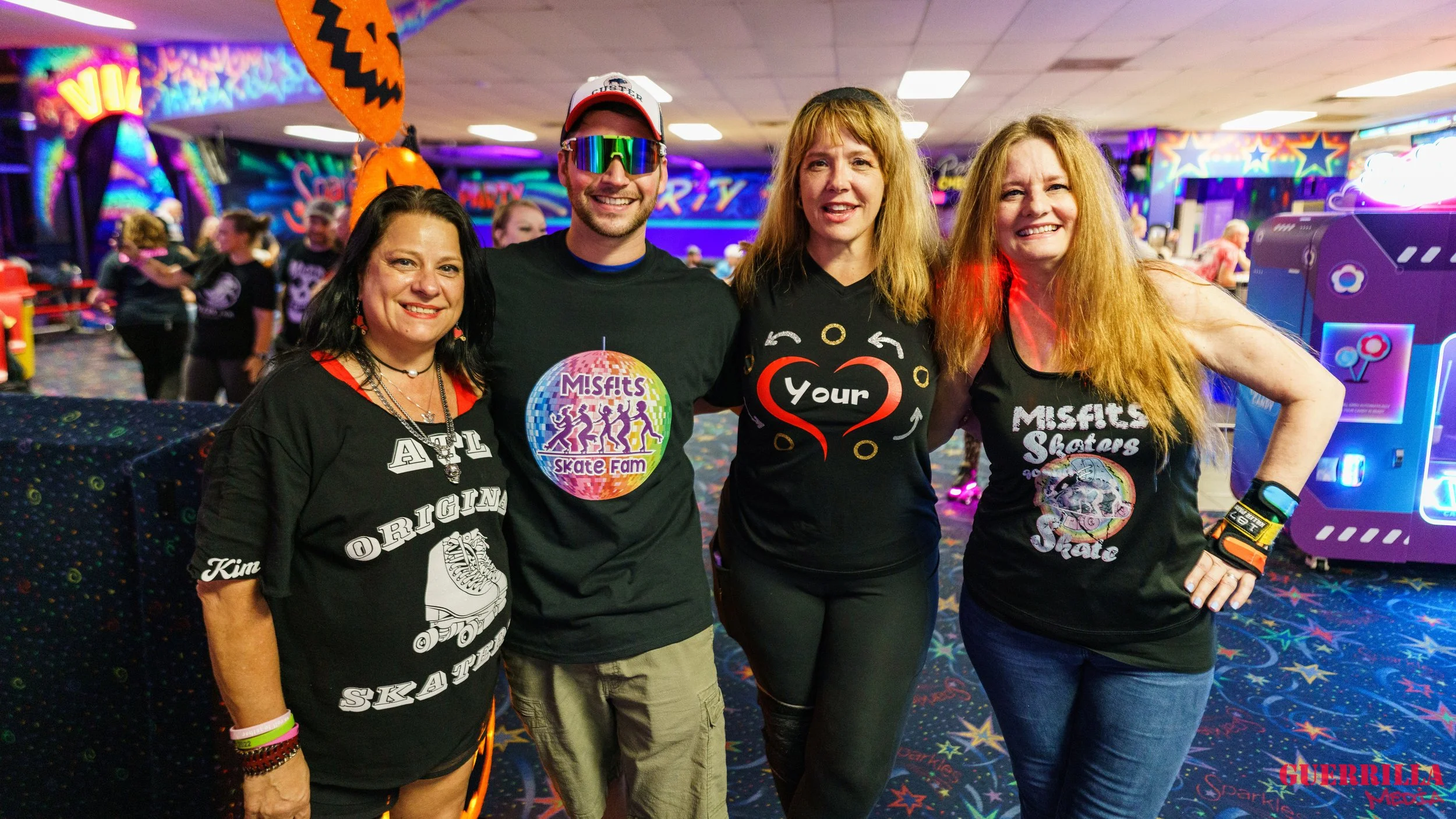 Four people posing together at a roller skating rink, all wearing black Misfits Skaters shirts, with a colorful neon background and arcade games in the background.