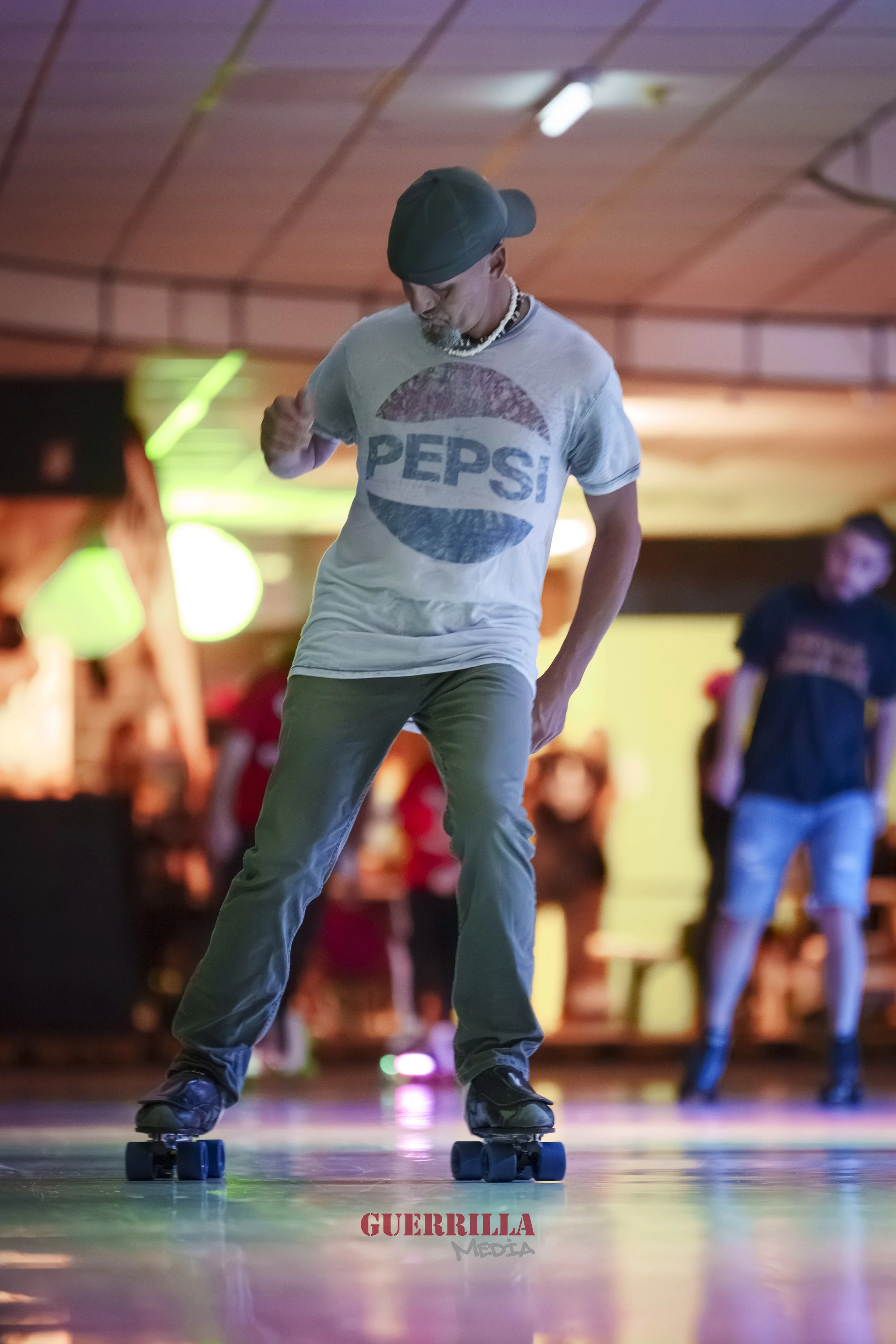 A person roller skating indoors, wearing a Pepsi logo t-shirt, a black cap, and dark pants with other people visible in the background.
