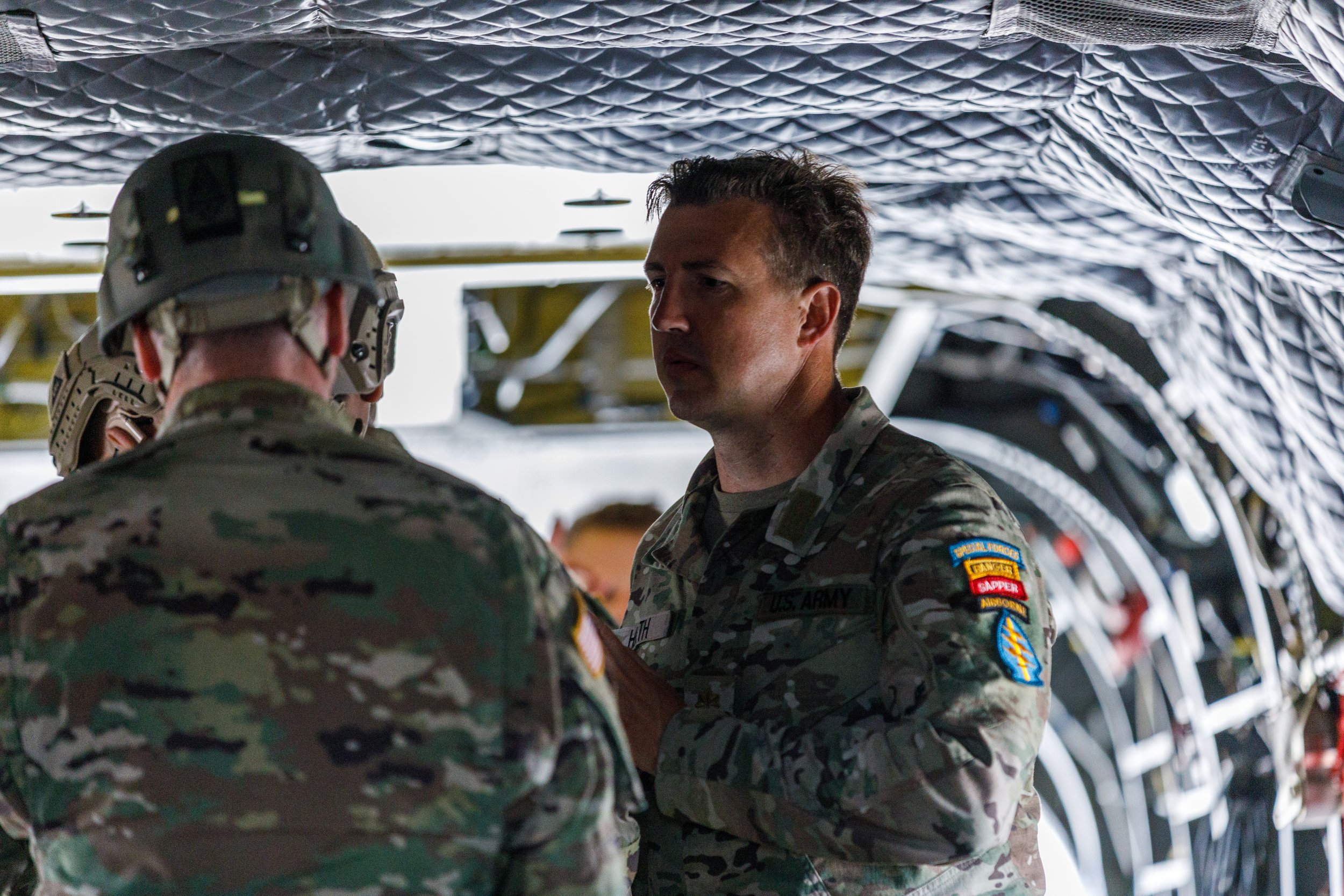 U.S. Army soldiers inside a military aircraft, wearing camouflage uniforms and helmets, engaged in a discussion or briefing.