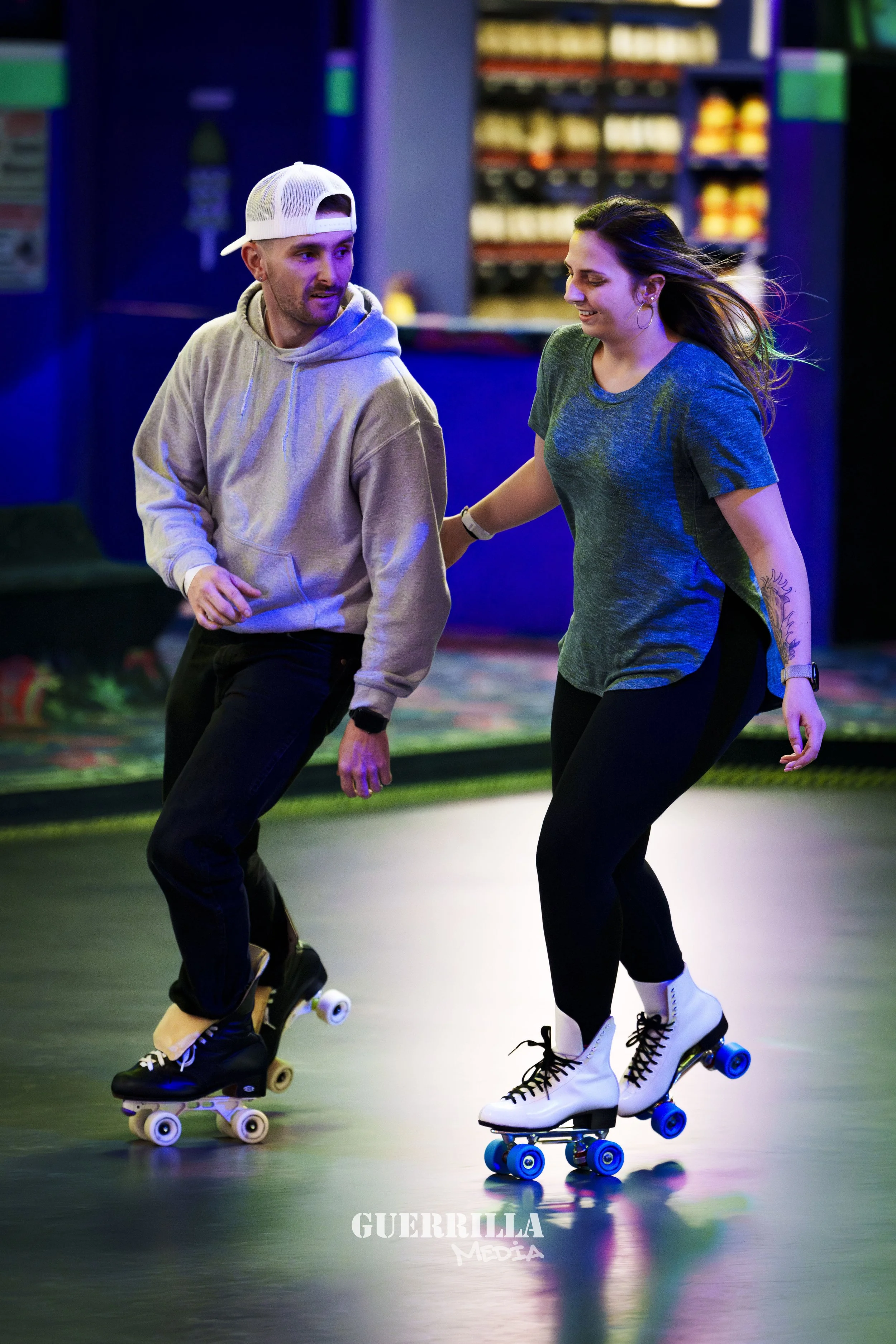 A man and woman roller skating indoors, smiling and enjoying. The man is wearing a gray hoodie, black pants, a white cap backwards, and black skates. The woman is wearing a blue t-shirt, black pants, white skates, and has a tattoo on her left forearm