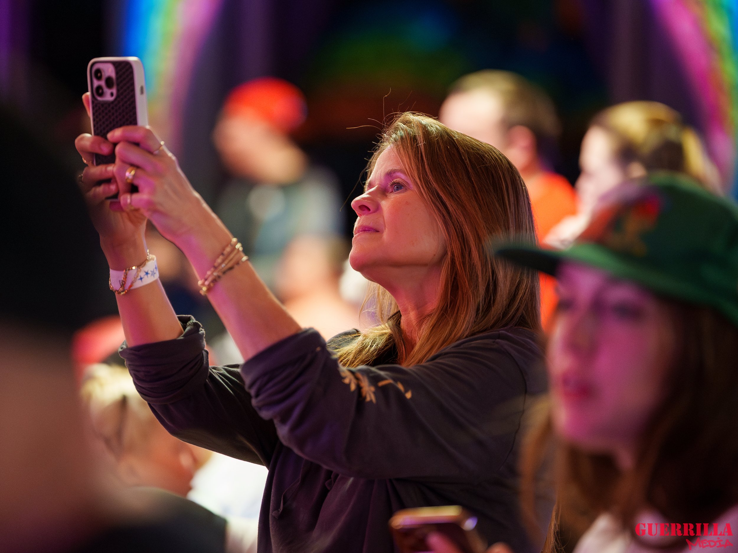 A woman with red hair taking a photo or video with her smartphone at an event, with colorful lights in the background and a woman wearing a hat in front of her.