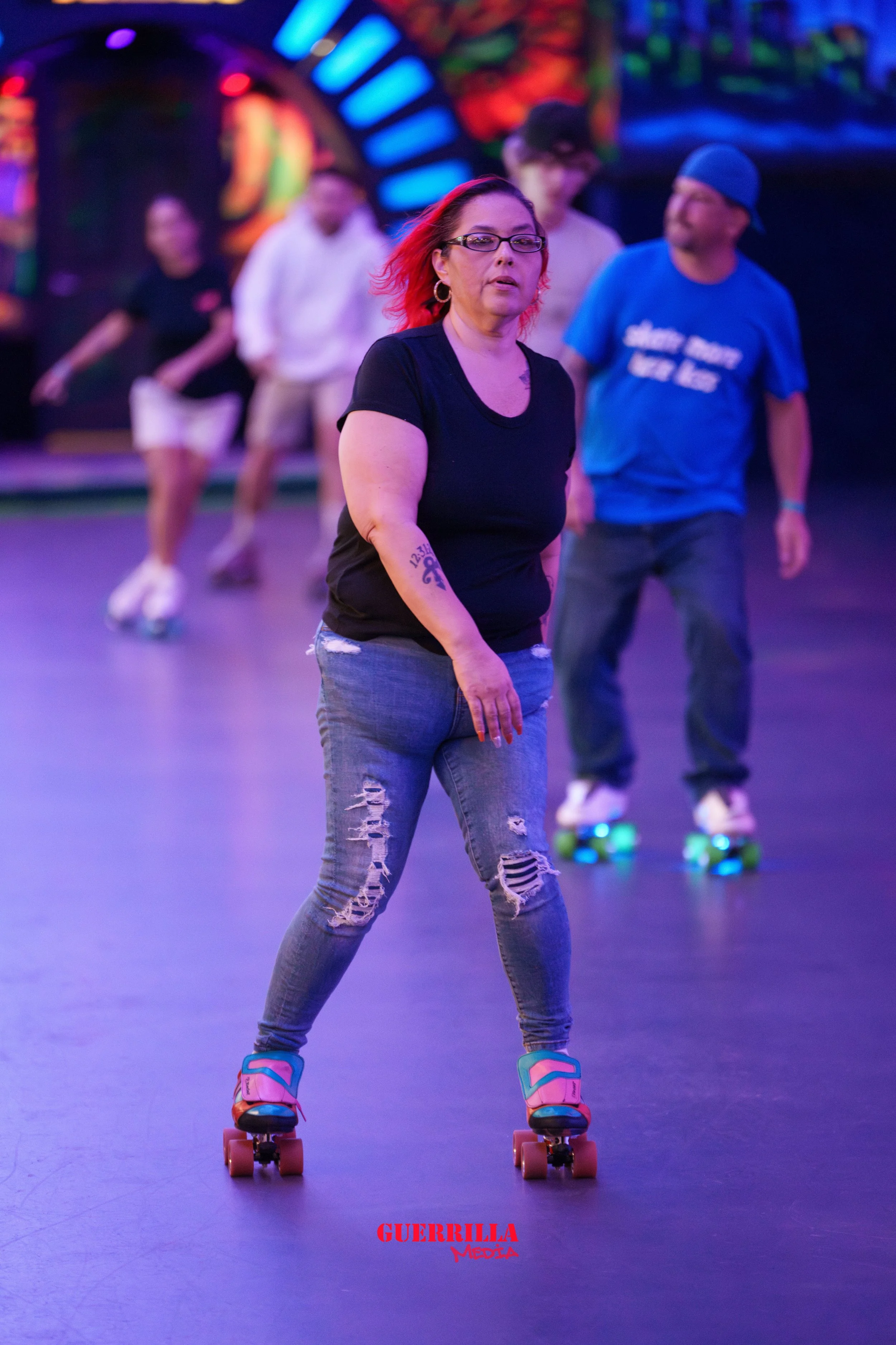 A woman with red hair and glasses skating at an indoor roller rink with colorful lights, wearing a black t-shirt and ripped jeans.