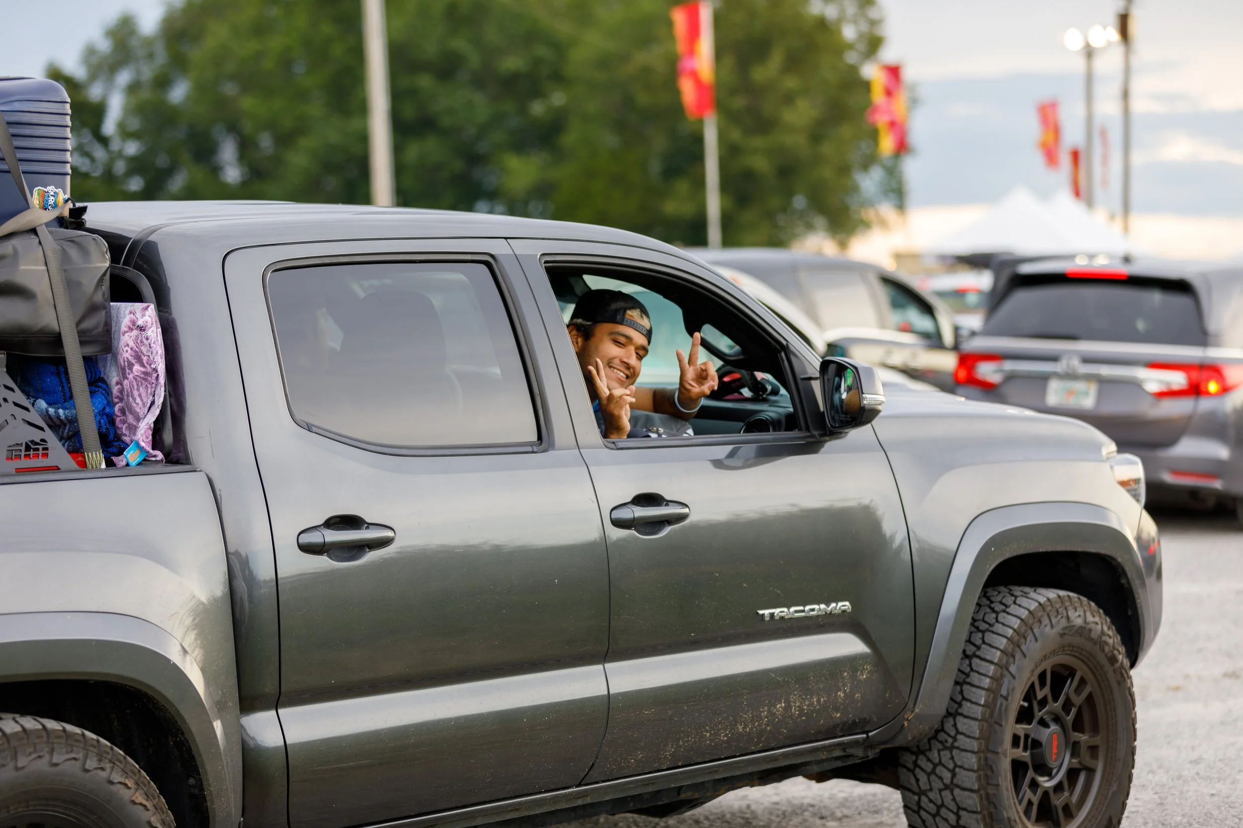 A young man smiling and making a hand gesture from the driver's side window of a gray Toyota Tacoma pickup truck, parked in a lot with other vehicles during daytime.