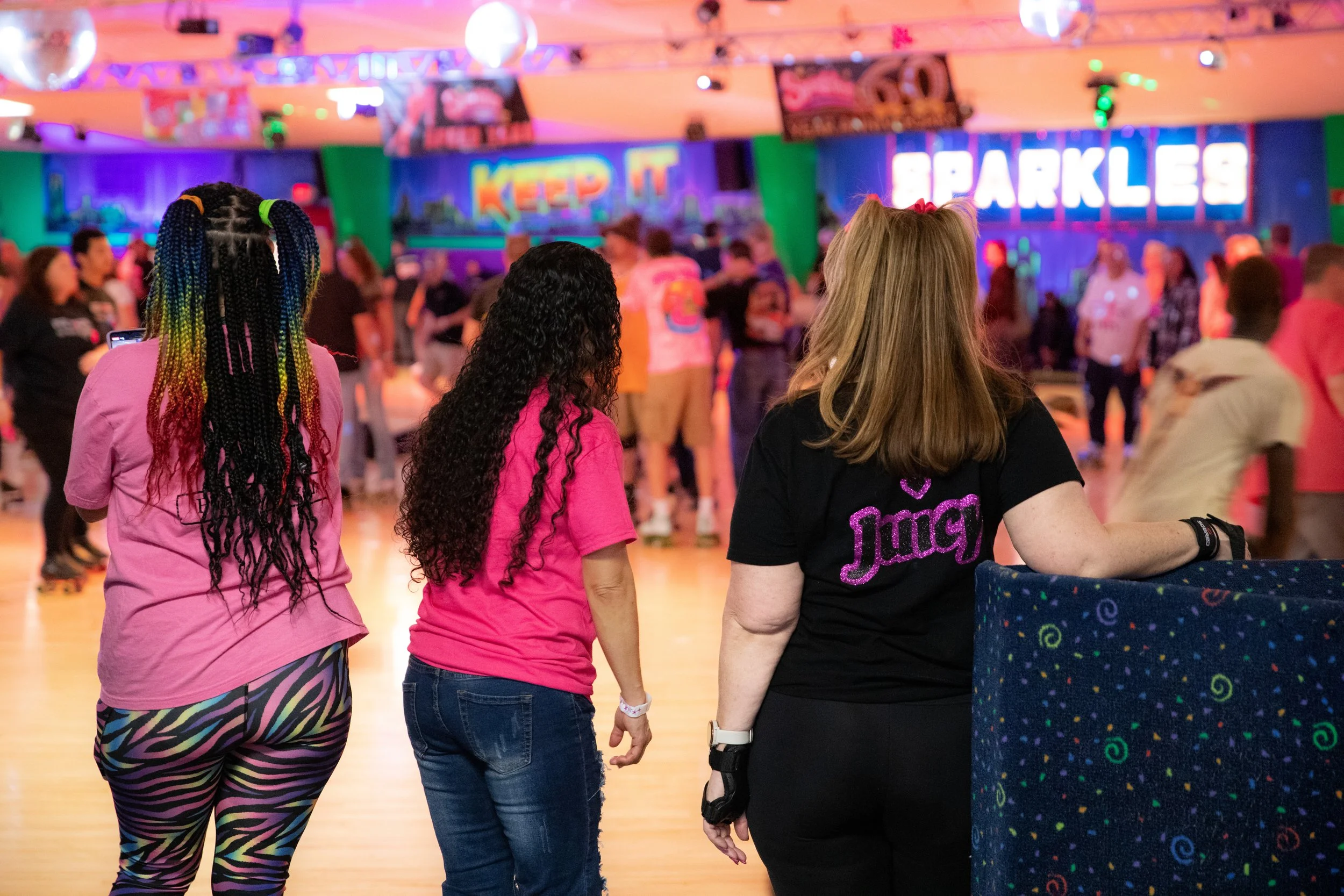 Three women watching people roller skating at an indoor roller rink with colorful neon signs and decorations.