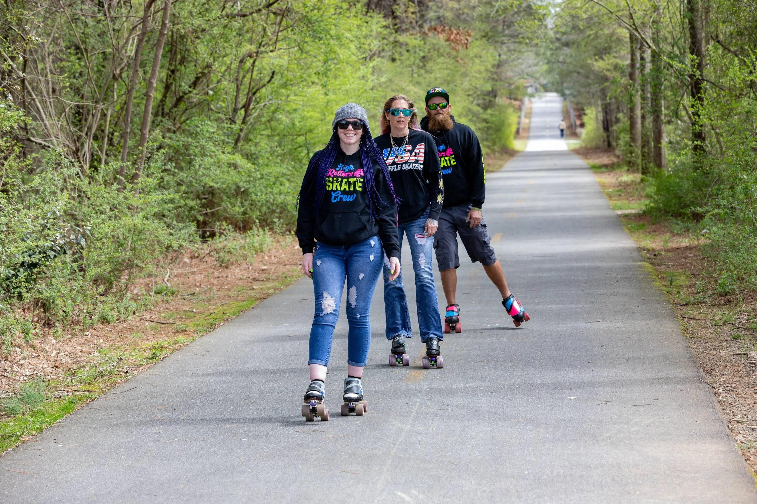 Three people roller skating on a paved trail through a green forest.