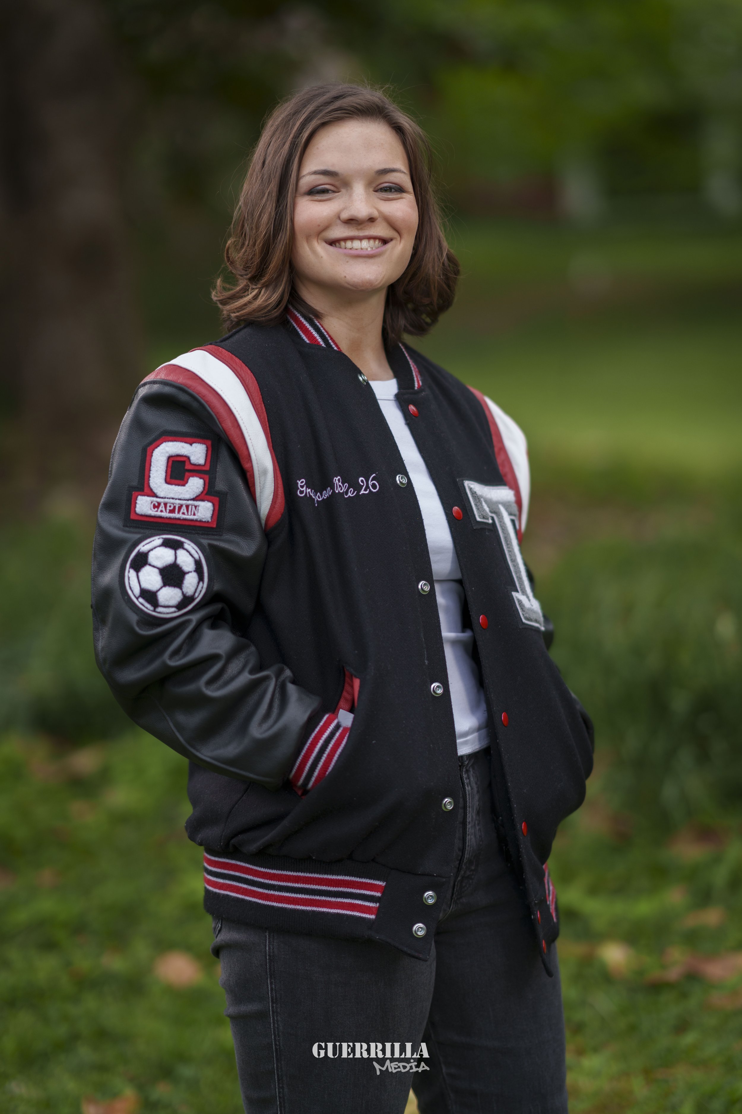 Young woman smiling outdoors, wearing a black and red varsity jacket with patches including a soccer ball, a letter 'T', a captain patch, and embroidered name and age, standing on green grass with blurred trees in background.