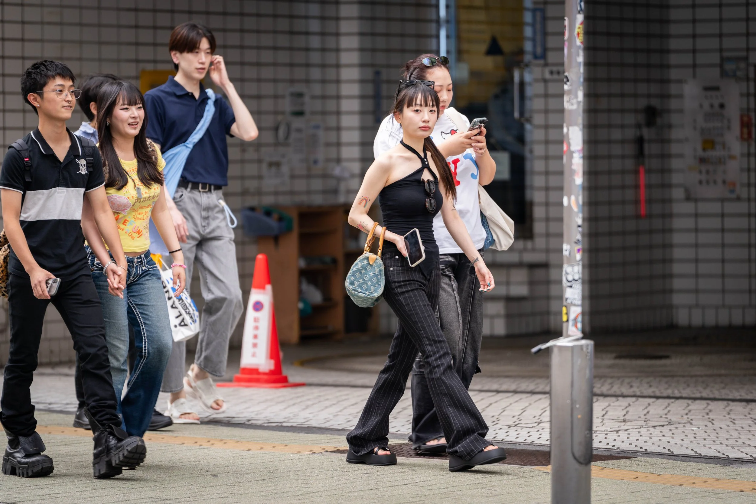 Group of young people crossing the street, some holding hands, with a woman in black pants and tank top in the foreground looking at her phone.