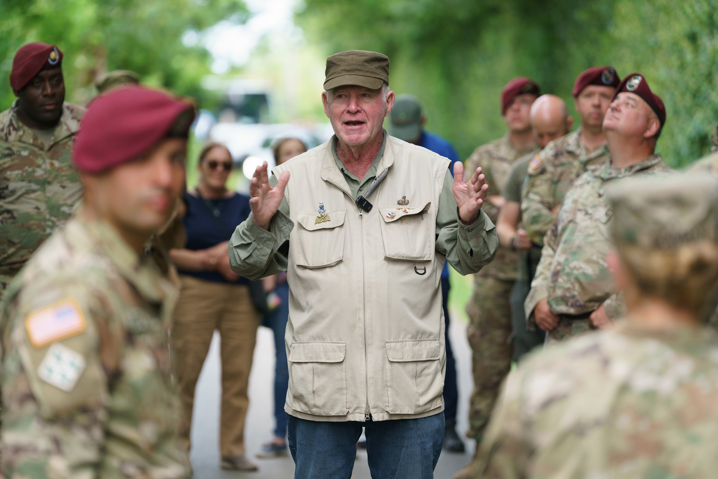 A man in a beige vest and cap speaking to military personnel in camouflage uniforms, some wearing maroon berets, outdoors in a forested area.