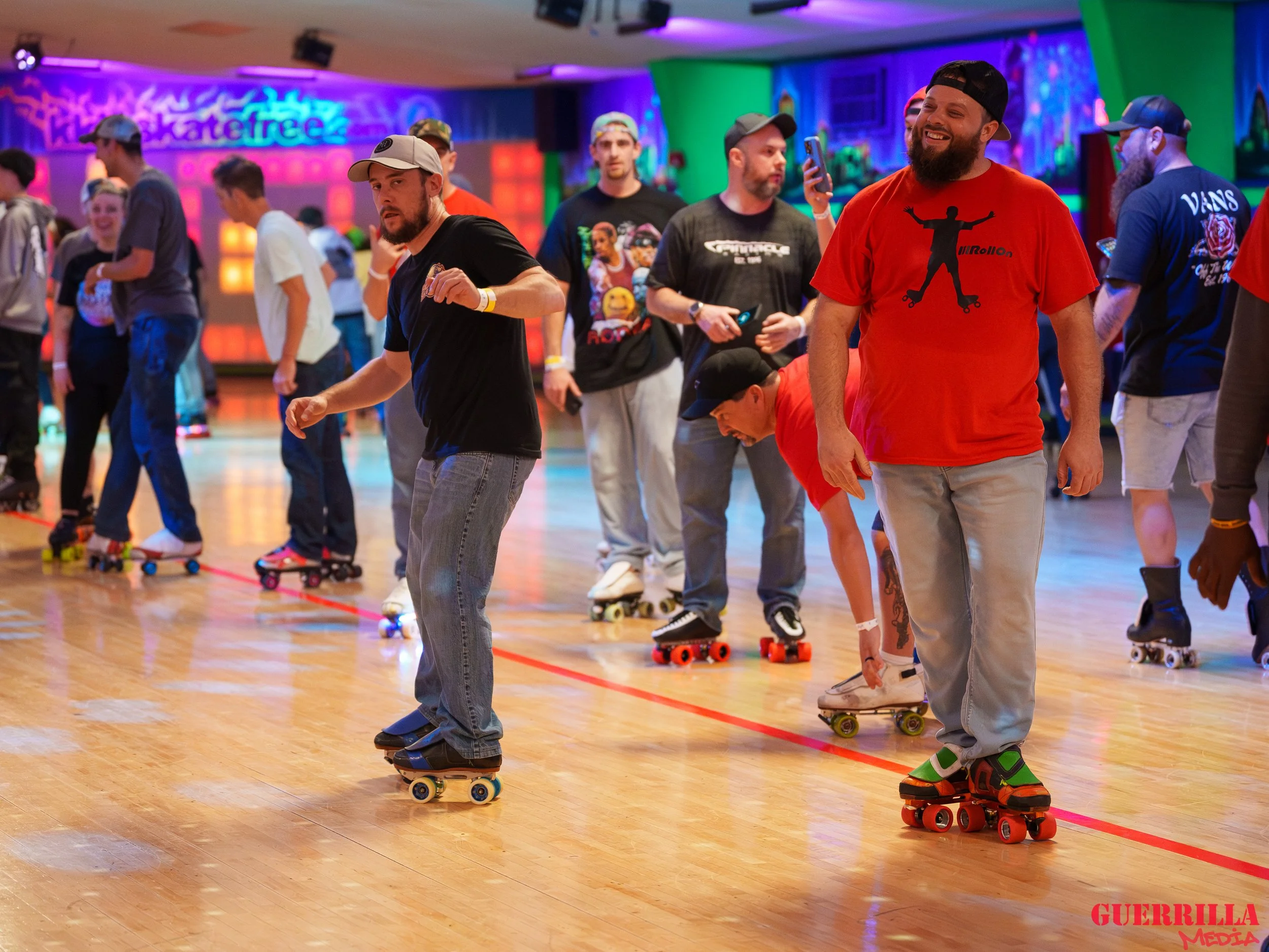 People roller skating in an indoor skate rink with colorful neon lights and graffiti-style wall art in the background. Some skaters are smiling and taking photos.