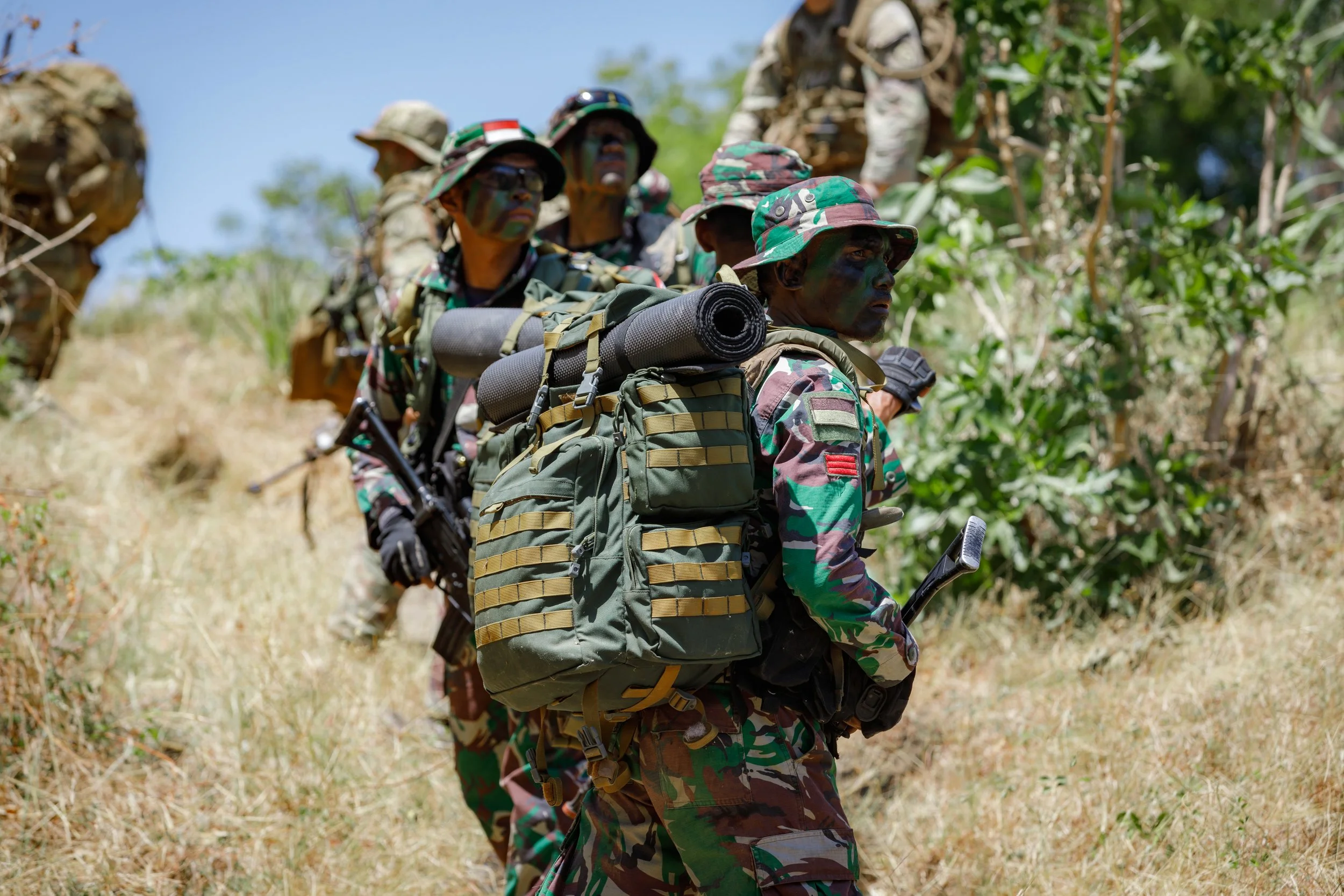 Group of soldiers in camouflage uniforms walking through a grassy area with trees, carrying backpacks and gear, on a sunny day.