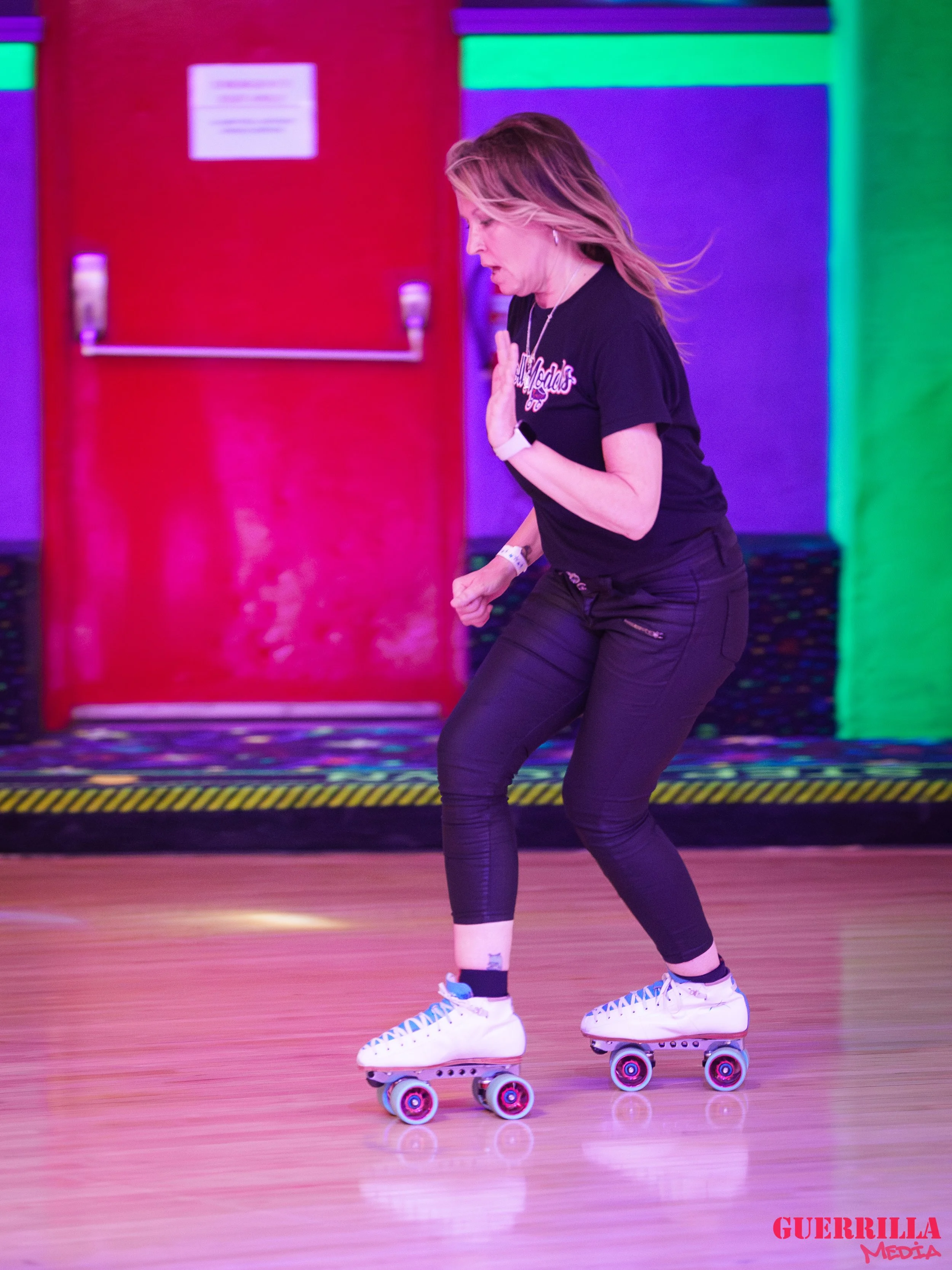 A woman roller skating indoors, wearing a black t-shirt and black pants, with her hands near her chest in a balancing pose, and a colorful background with a red door and multicolored lighting.