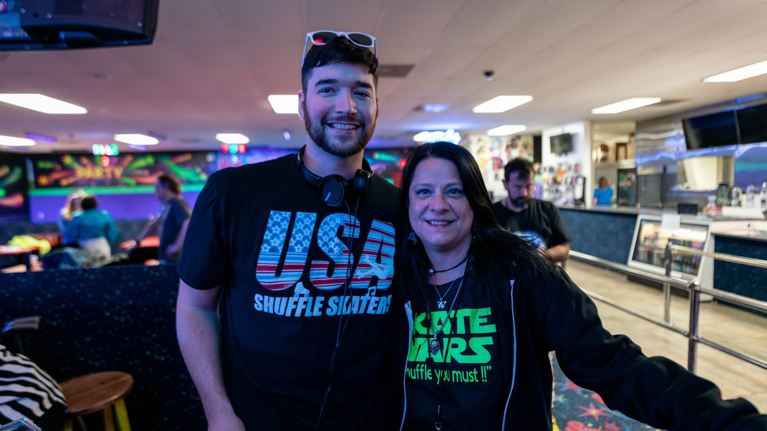 Two people at a neon-lit indoor bowling alley, smiling at the camera. The man on the left is wearing a black T-shirt with an American flag and USA written on it, along with sunglasses on his head and headphones around his neck. The woman on the right
